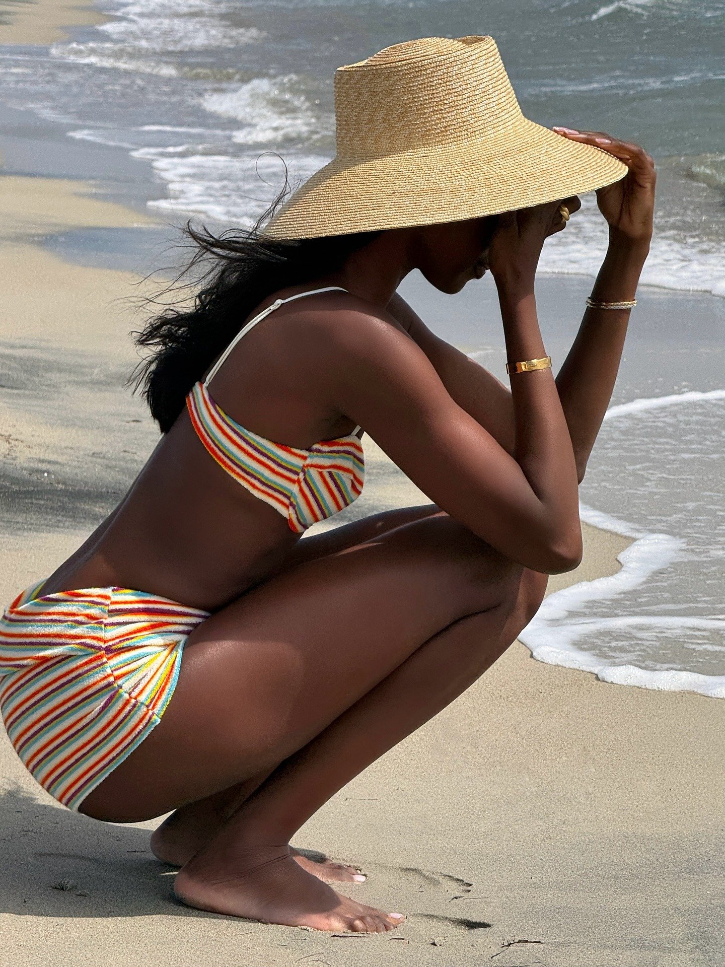 A imagem retrata uma mulher na praia, usando um chapéu de palha amplo, perfeito para proteção solar. Ela veste um biquíni listrado, que destaca as cores vibrantes da estação. A pose, com um toque de descontração, reflete as tendências de chapéu, que são e