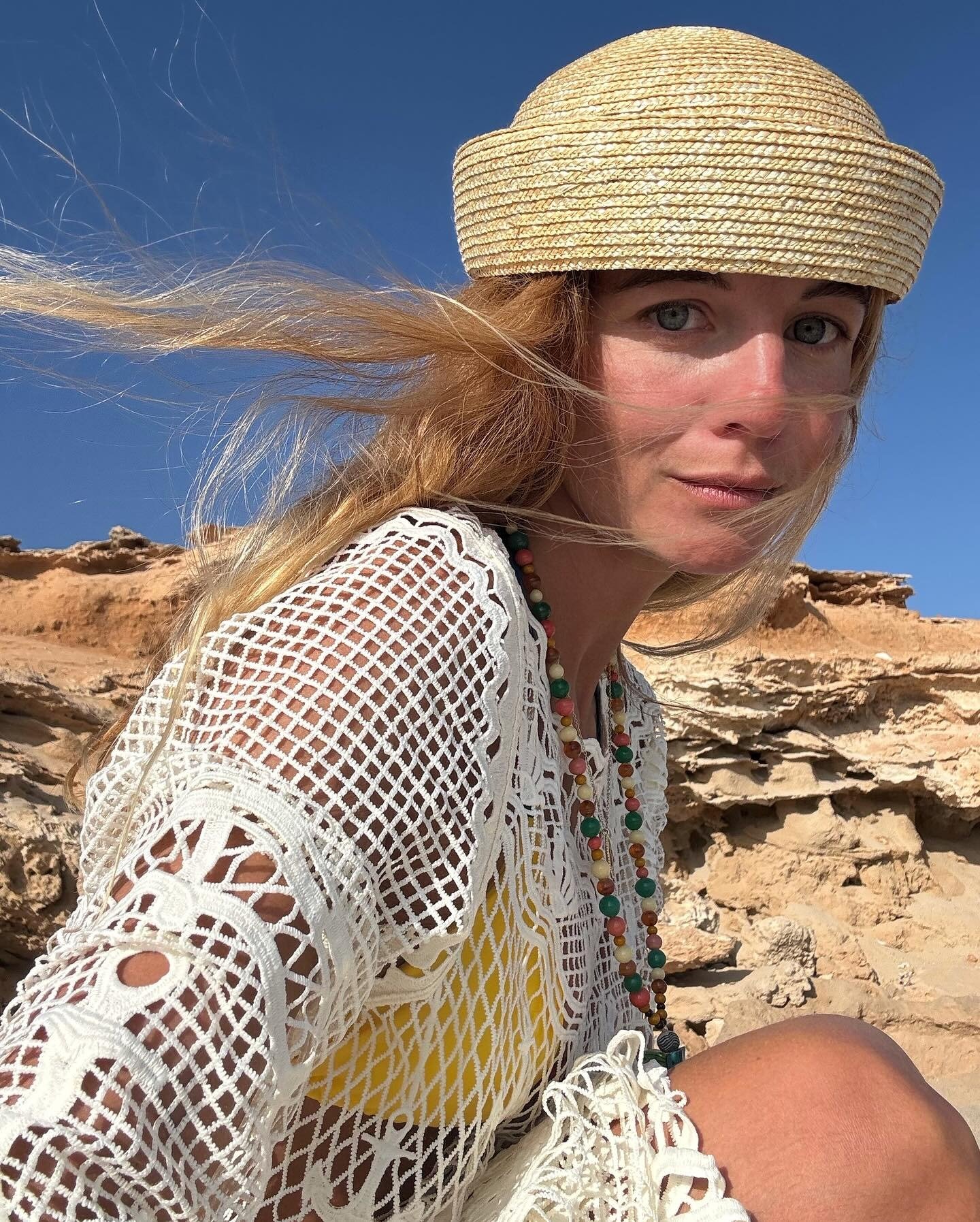 A imagem apresenta uma mulher usando um chapéu de palha, que reflete as tendências de chapéu atuais na moda. Seu look é complementado por um kimono de crochê branco e um colar colorido, evocando um estilo boho-chic. O fundo natural destaca a leveza e a el