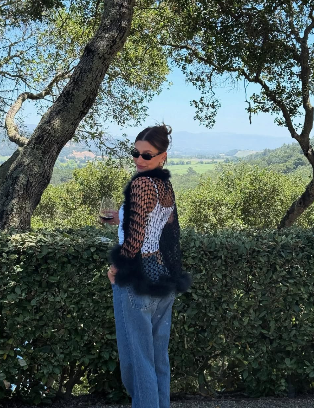 Hailey Bieber em vestes casuais com um copo de vinho, posando ao lado de uma cerca e árvores com uma paisagem ao fundo.