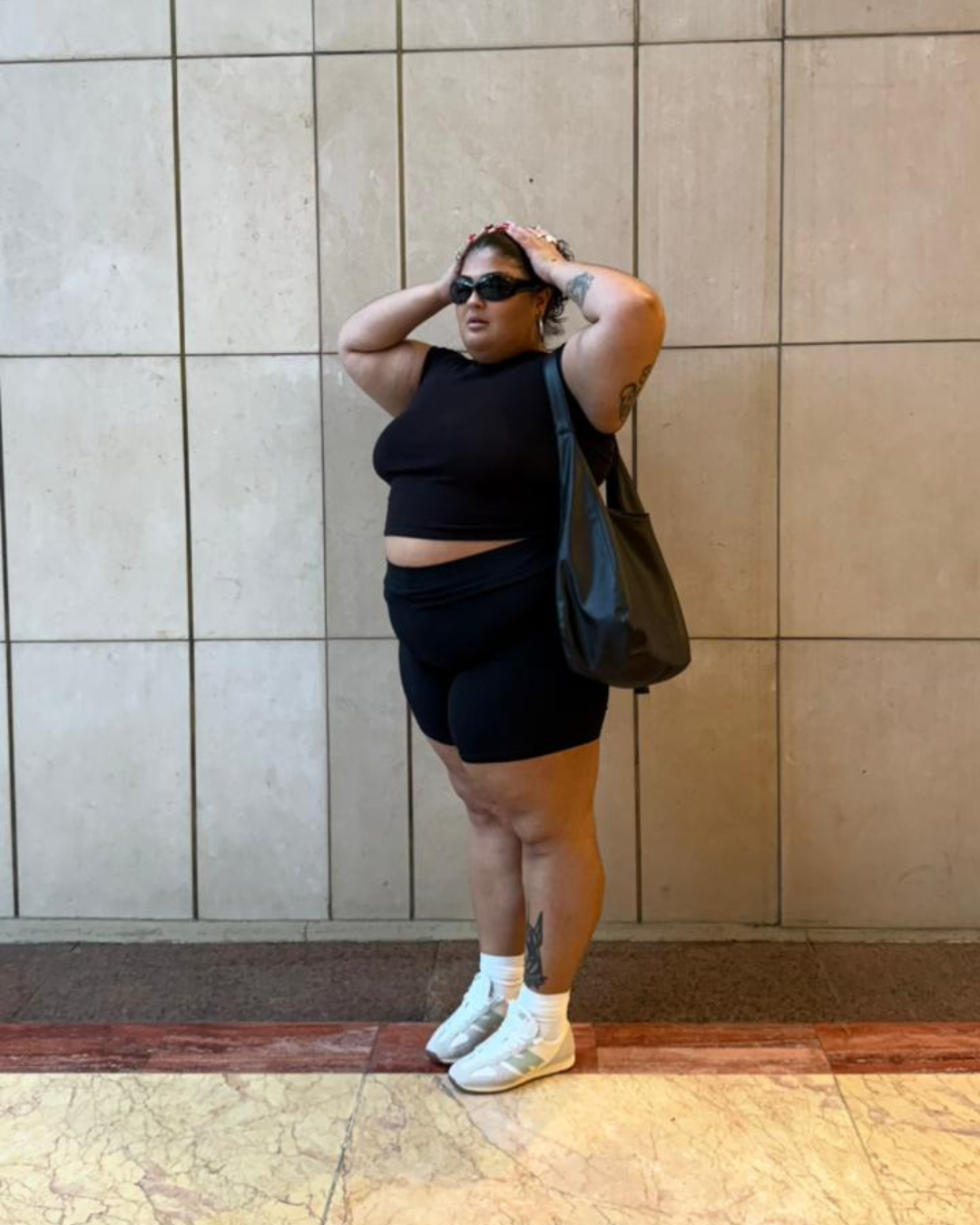 Pessoa com roupa preta e óculos escuros apoia as mãos na cabeça. Veste tênis casual branco.