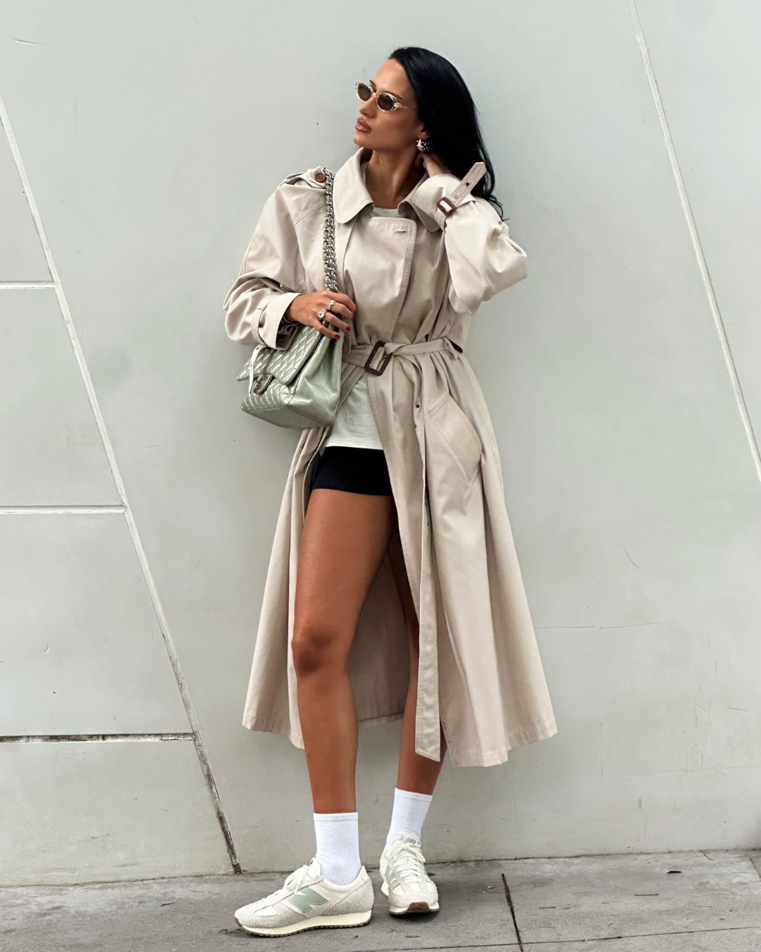 Pessoa em trench coat bege com tênis casual branco, óculos de sol e bolsa prateada, em frente a parede clara.