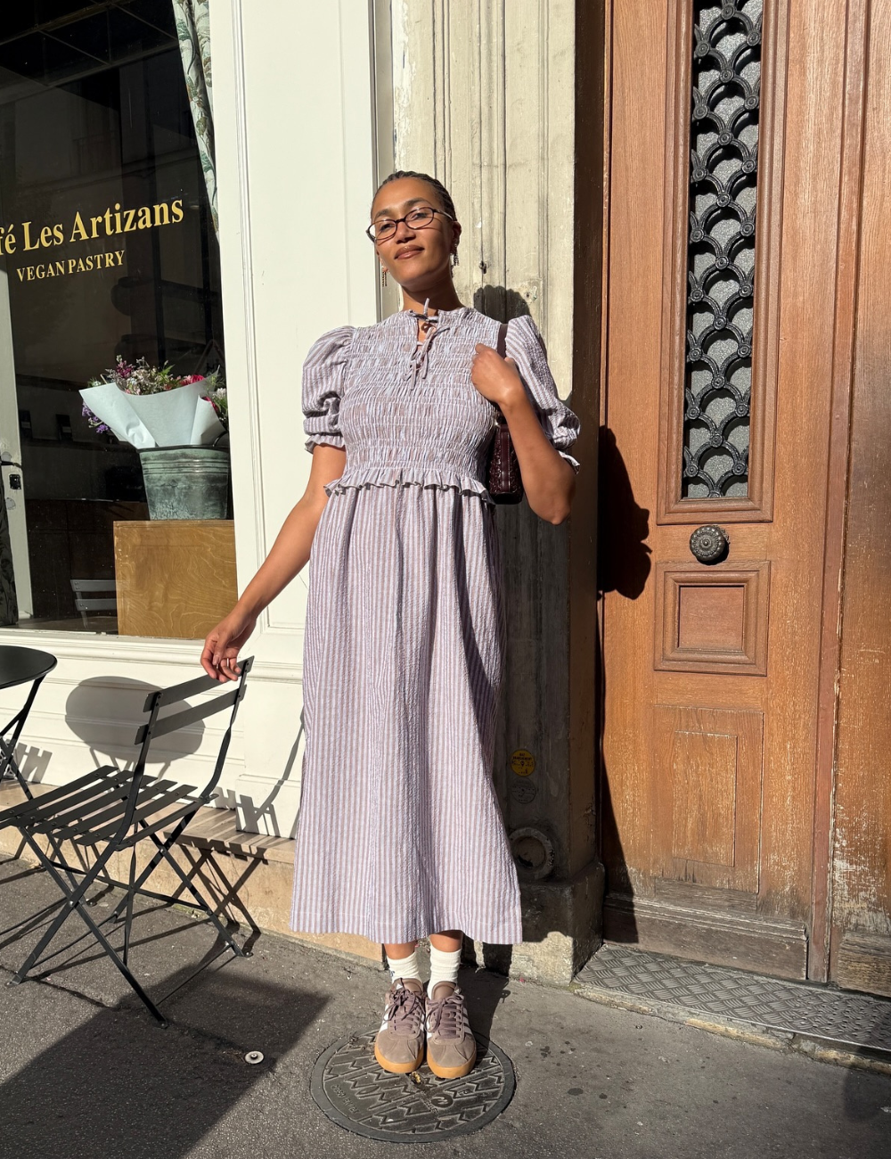Pessoa em vestido listrado, óculos, em frente a um café. Usa tênis confortável feminino em tom neutro.