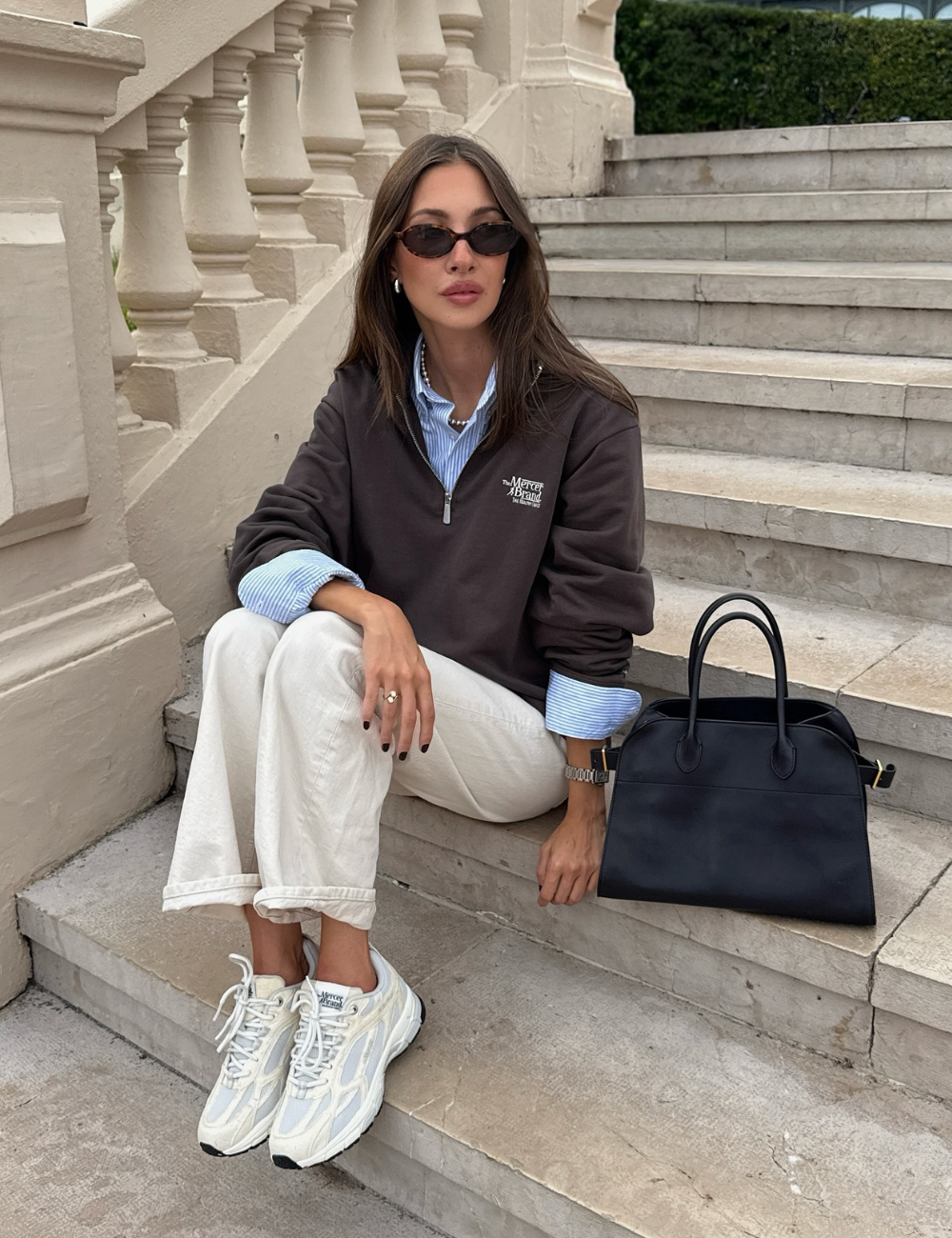 Uma mulher com calça branca e blusa escura usa tênis confortável feminino, sentada em escadas ao lado de bolsa preta.
