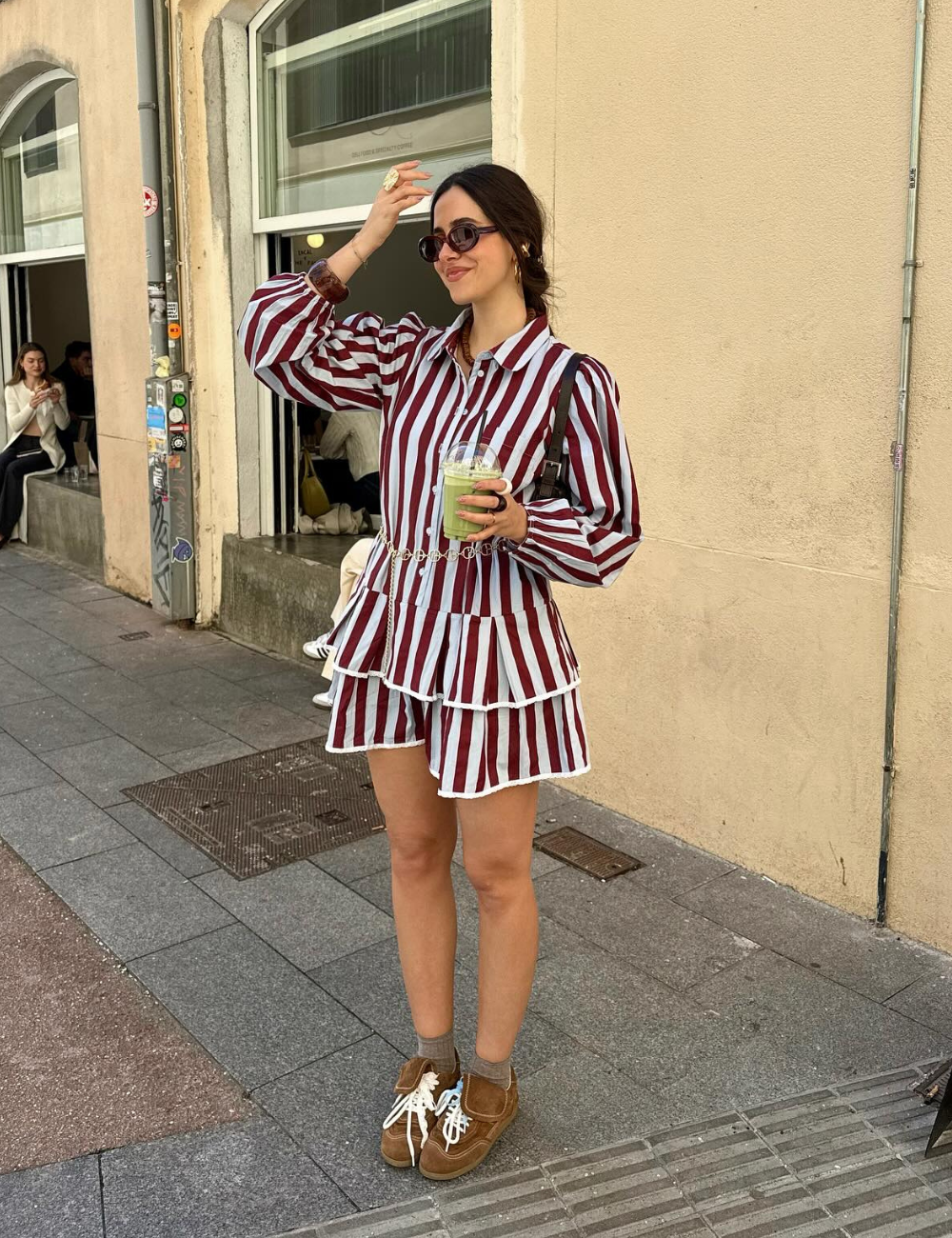 Pessoa usando vestido listrado em vermelho e branco, óculos escuros, segurando bebida e calçando tênis marrons.