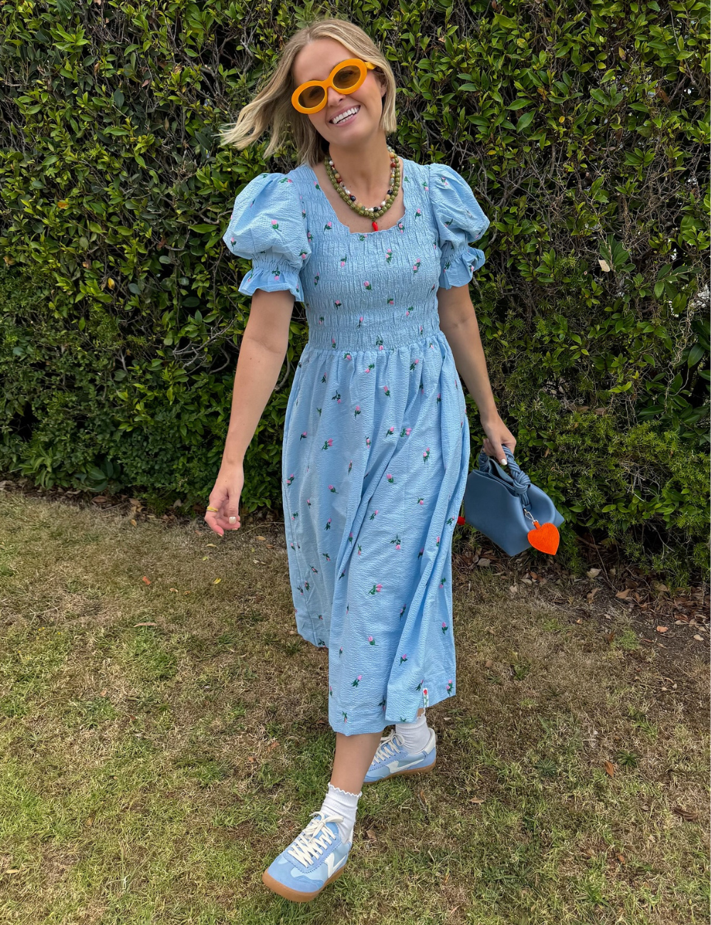 Vestido azul estampado usado com tênis e óculos laranja, em frente a um arbusto verde.