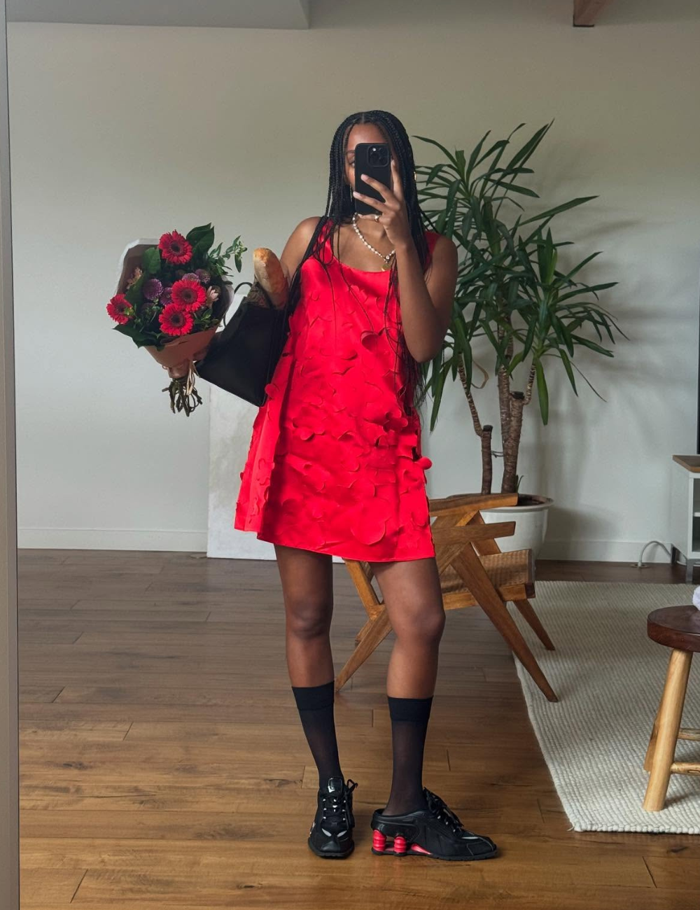 Pessoa com vestido vermelho, tênis preto e meias, segurando flores e baguete em ambiente interno.