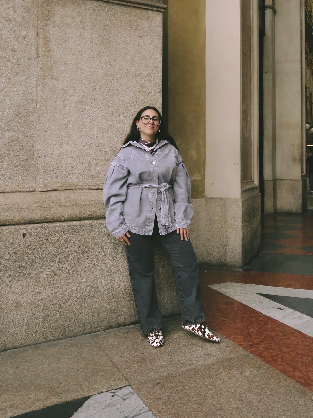 Jovem usando casaco jeans e sapatos estampados na Milão Fashion Week, encostada em parede.