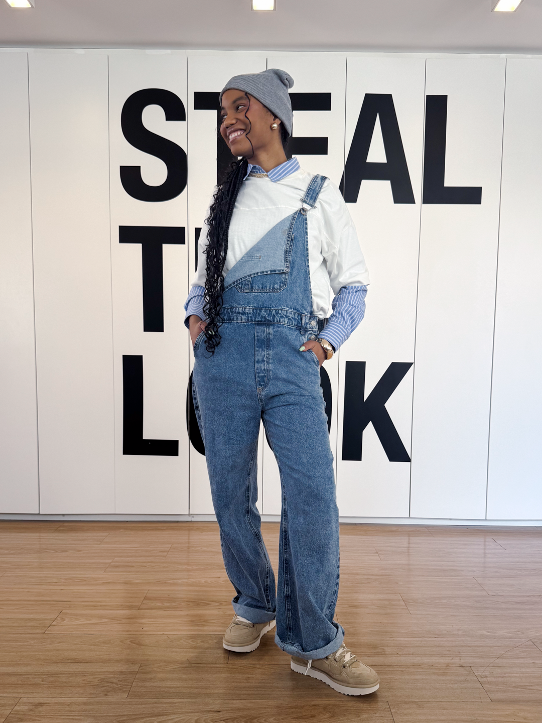 A imagem mostra uma jovem estilosa usando um macacão jeans oversized, combinado com uma camiseta branca e uma camisa de manga longa listrada. Os tênis de camurça em tom bege complementam o look casual e moderno, trazendo conforto e sofisticação. O cenário