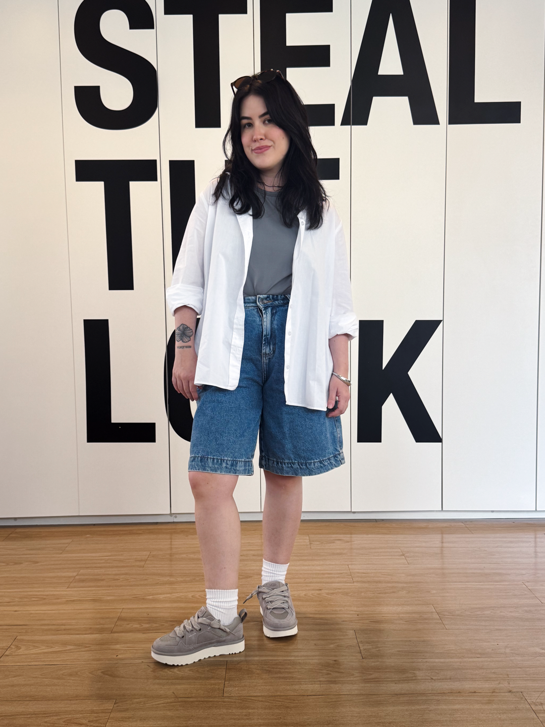 A imagem apresenta uma jovem usando um look casual e moderno, com destaque para os tênis de camurça cinza que conferem conforto e estilo ao visual. Ela combina uma camiseta cinza com uma camisa branca oversized e shorts jeans. Os detalhes do conjunto, com