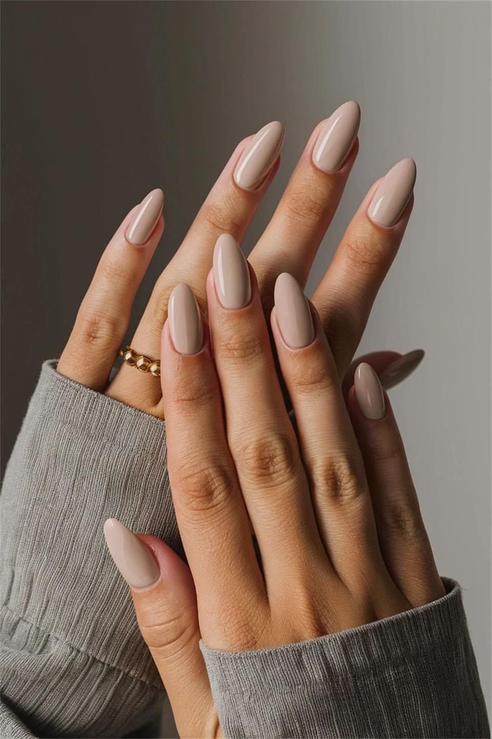 ideias de unhas nude alongadas e lisas, mãos cruzadas, detalhes de anel dourado e manga de suéter cinza.