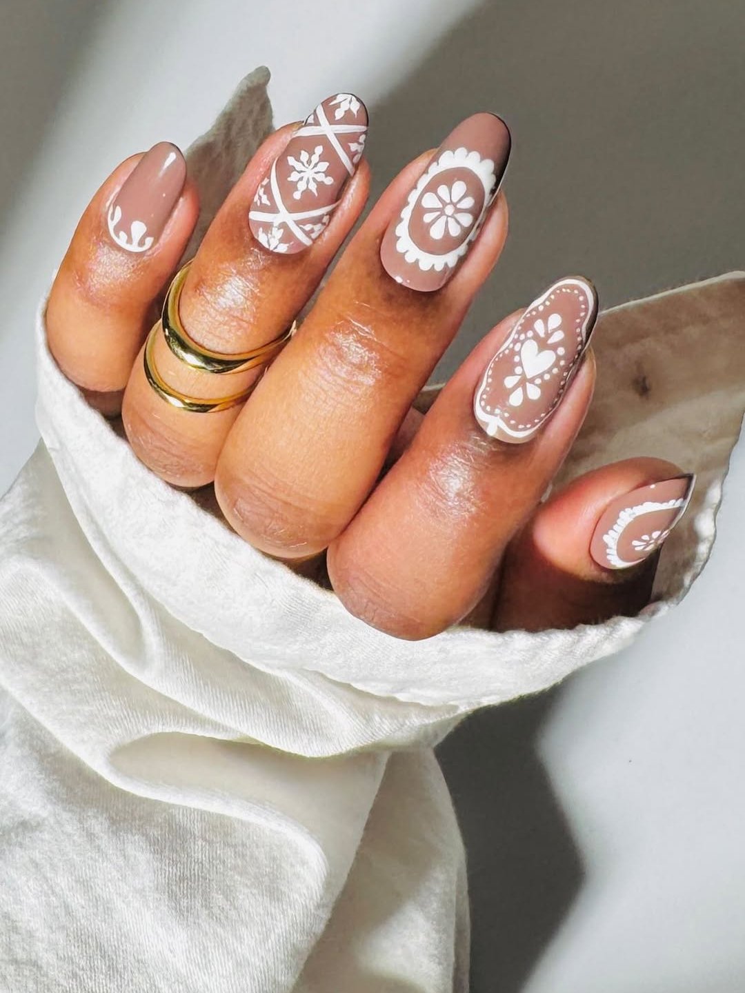 Ideias de unhas nude decoradas com detalhes brancos em formatos geométricos e florais. Anéis dourados com fundo em tecido branco.