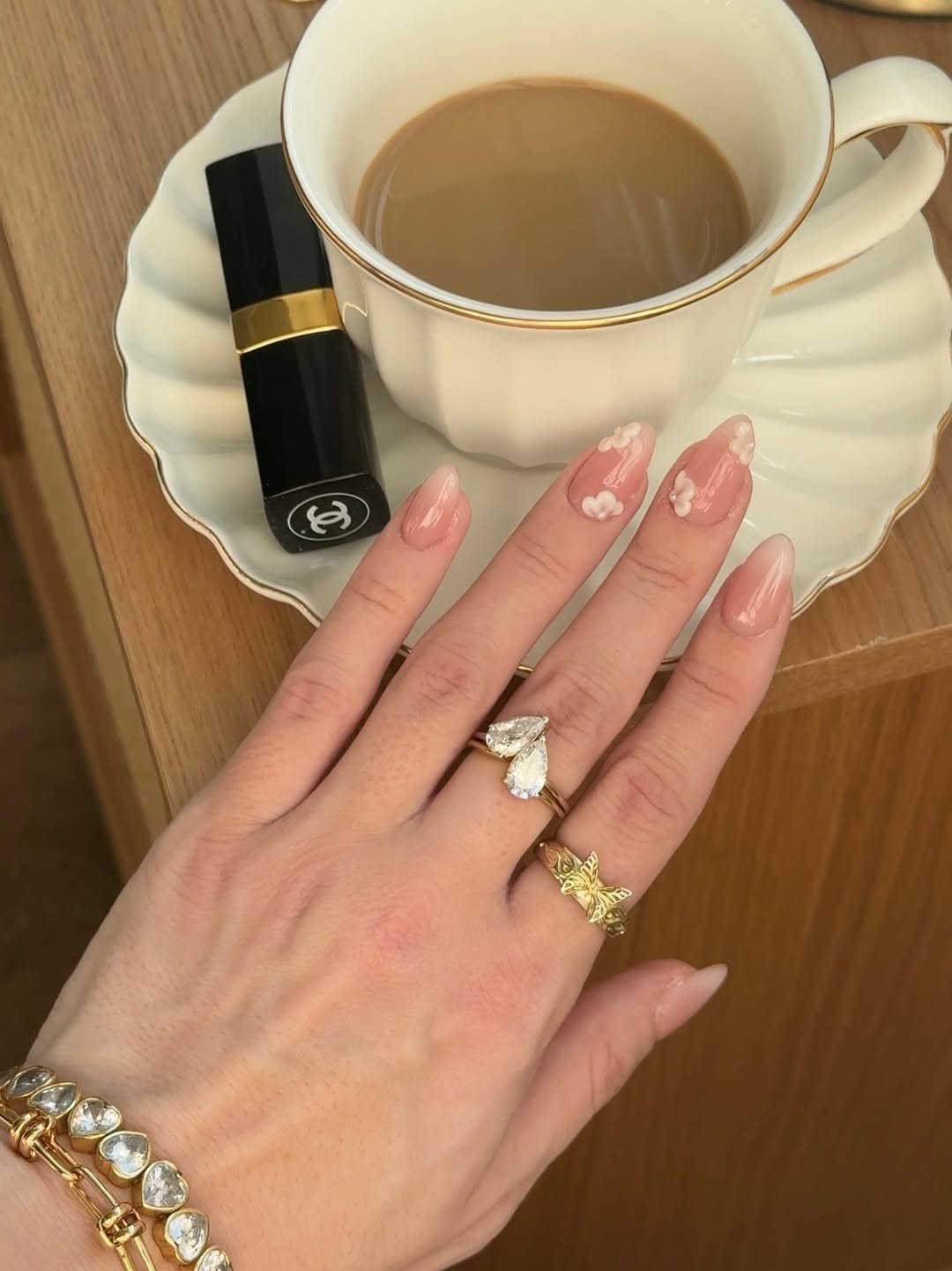 Ideias de unhas nude com detalhes florais delicados, acompanhadas por anéis elegantes, ao lado de um café e batom preto.