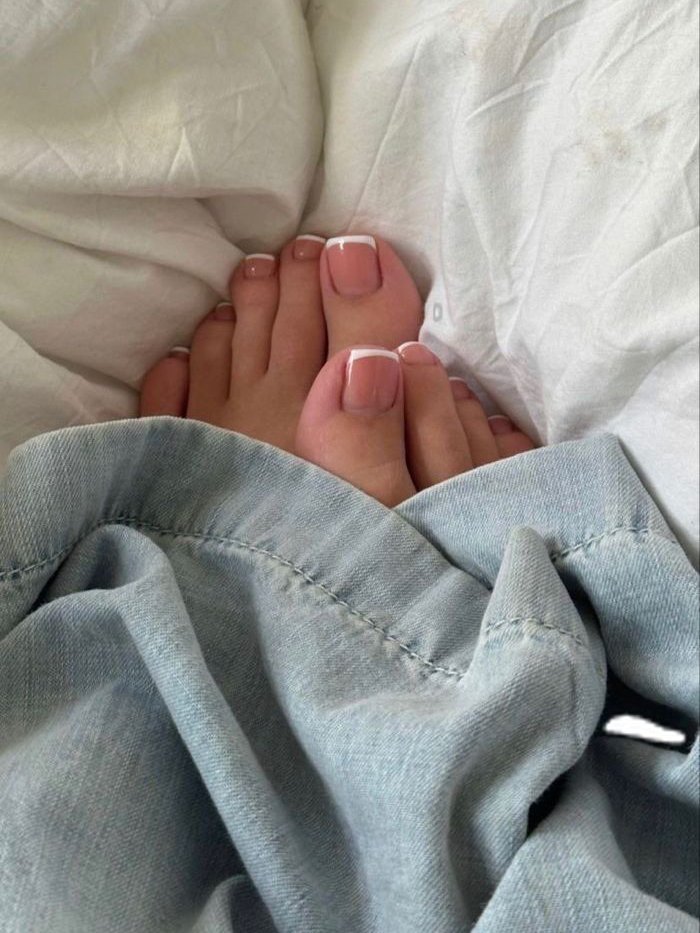 Unhas dos pés com esmalte nude decorado com ponta branca, sobre lençóis brancos e envolvendo jeans.