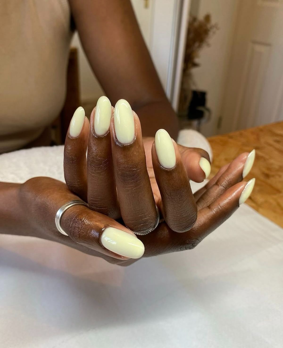 Unhas básicas em tom amarelo claro destacam-se na imagem, com formato oval e acabamento brilhante. As mãos, de pele escura, exibem um cuidado elegante, complementadas por anéis prateados.