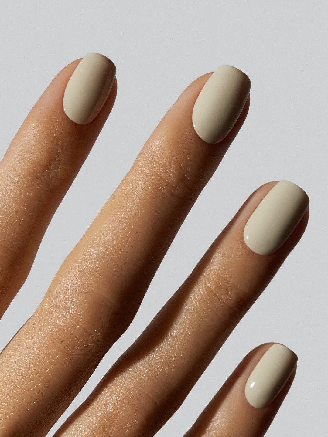 Unhas básicas em tom nude, bem cuidadas e com acabamento brilhante, destacam-se em um fundo neutro. A forma ovalada das unhas confere um toque elegante e moderno, ideal para diversas ocasiões.