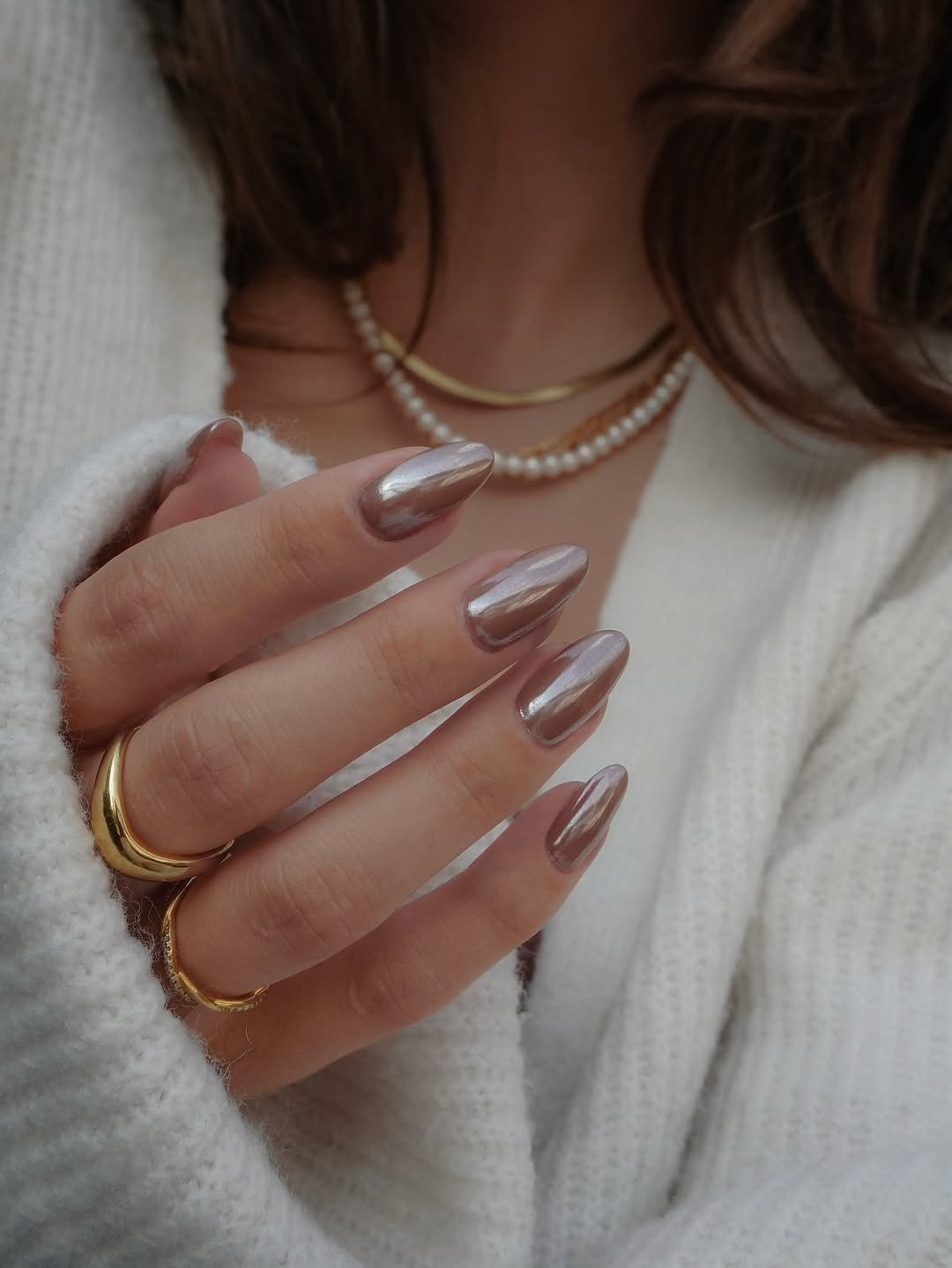 Unhas básicas em tom metálico prateado destacam-se na imagem, com formato ovalado e acabamento brilhante, conferindo elegância e sofisticação. A mão, adornada com anéis dourados, complementa o look.