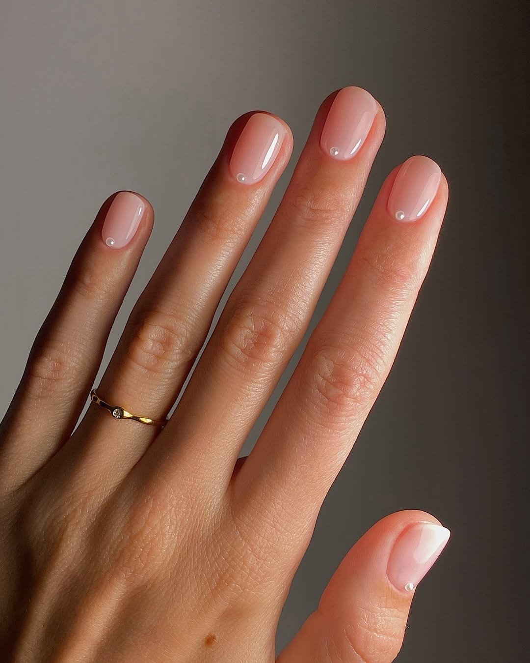 Unhas básicas em tom nude, com acabamento brilhante e delicadas pérolas na base, destacam a elegância e a simplicidade. A mão, com anéis finos de ouro, complementa o visual minimalista.