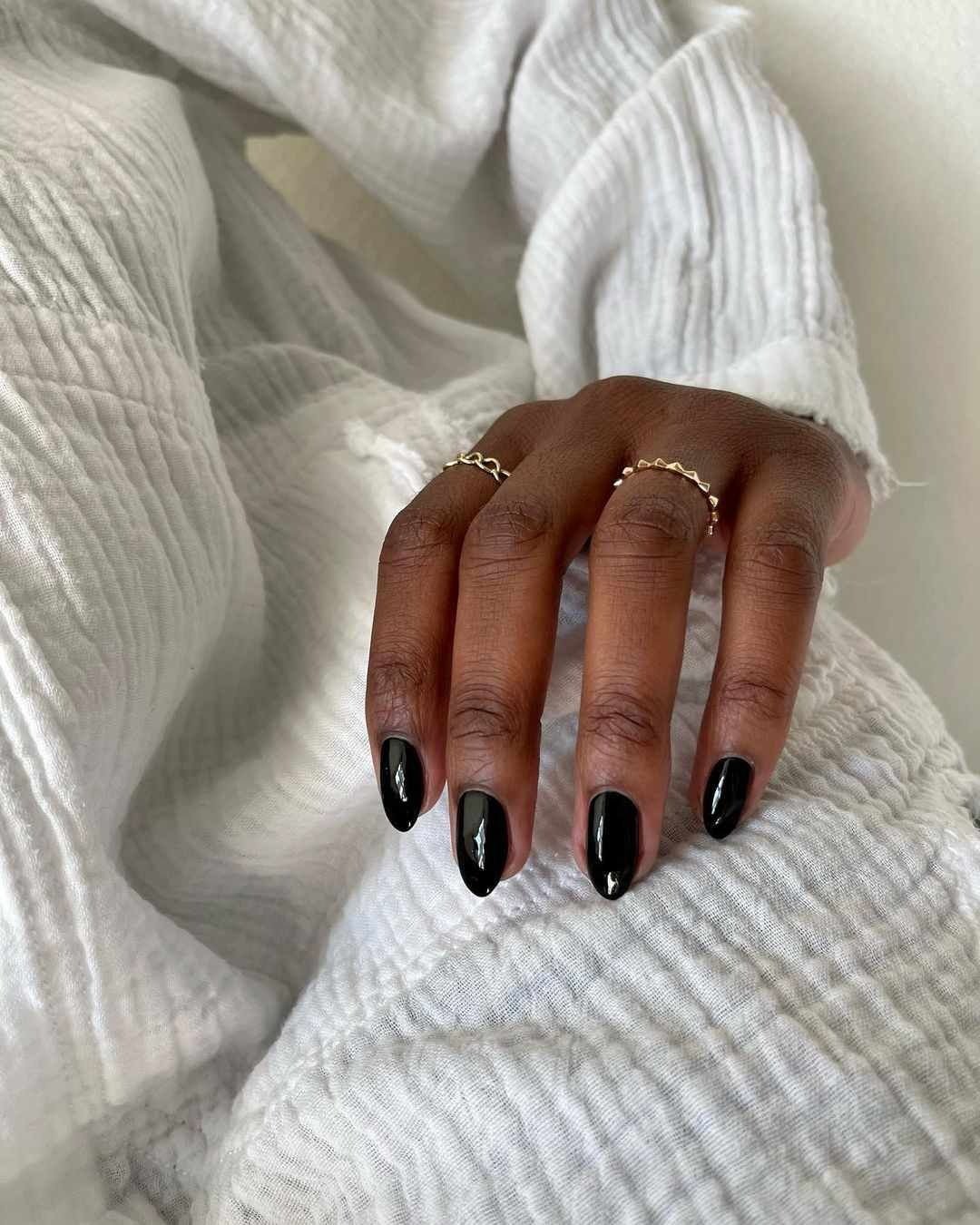 Unhas básicas em formato almond (amêndoa) estão pintadas de preto brilhante, destacando-se em contraste com a pele. A mão delicadamente repousa sobre um tecido branco texturizado, transmitindo elegância.