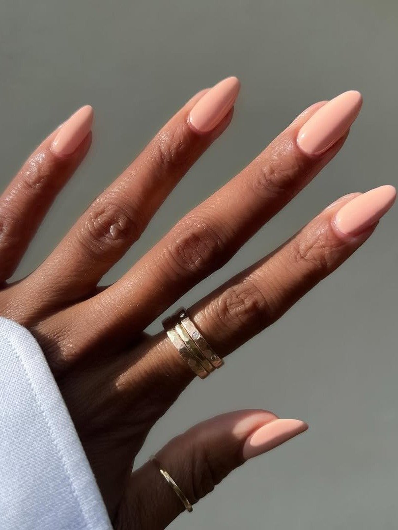 Unhas básicas em tom nude, com formato ovalado, destacam-se na imagem, transmitindo elegância e simplicidade. A mão é adornada com anéis dourados, complementando o visual.