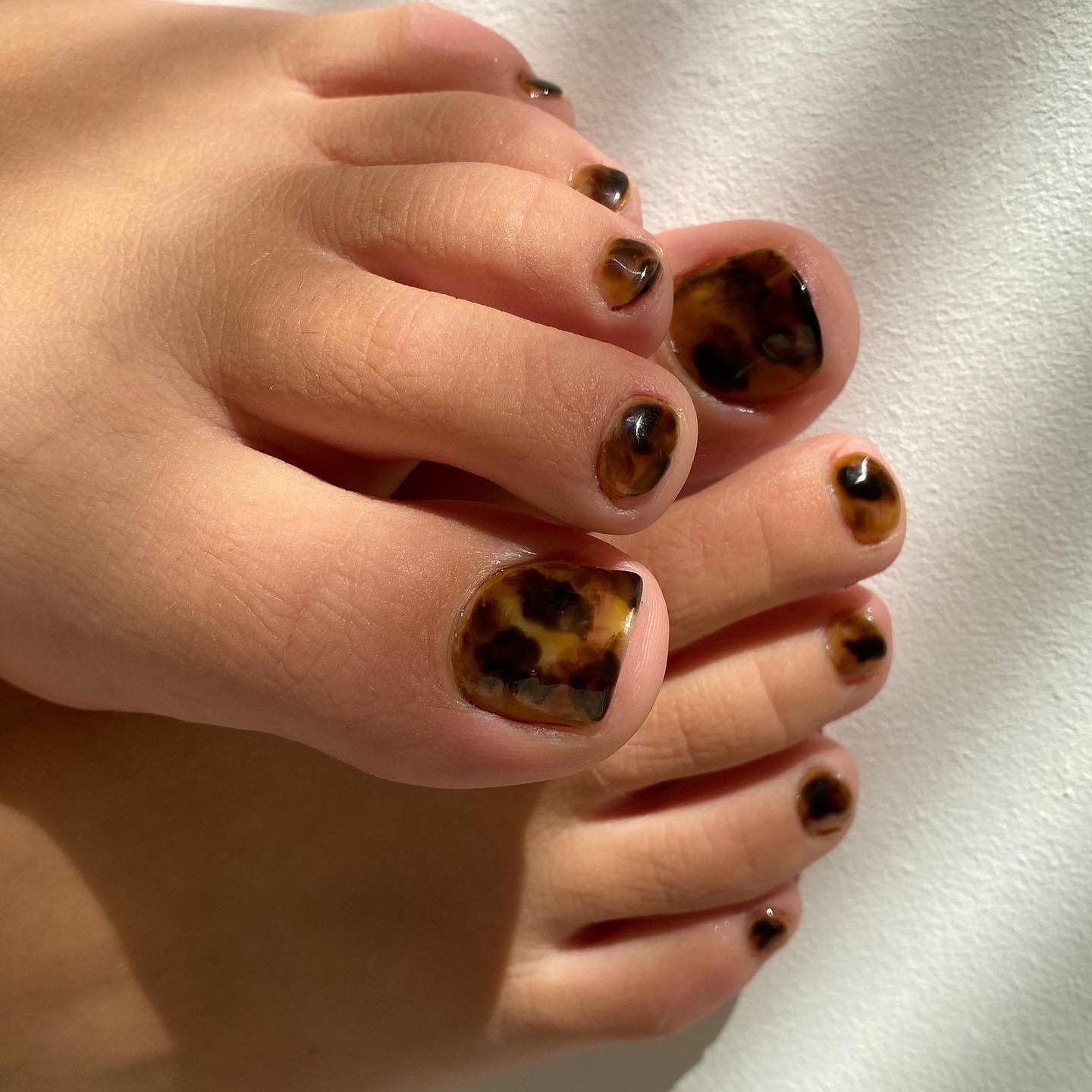As unhas decoradas do pé apresentam um design sofisticado em tons de marrom e preto, reminiscentes de padrão de tartaruga. Esse estilo é ideal para quem busca elegância e originalidade na pedicure. A combinação de cores quentes e a aplicação cuidadosa des