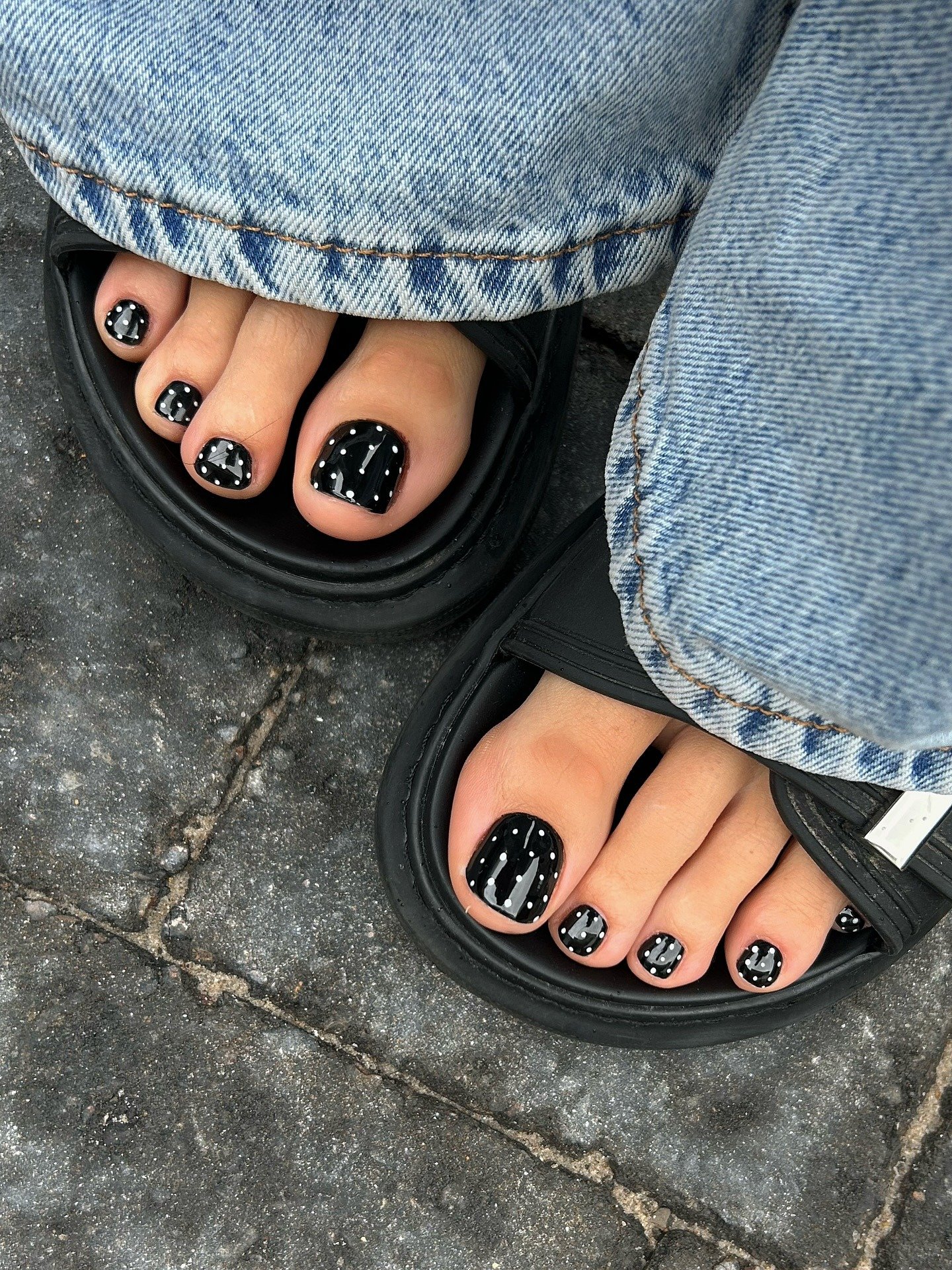 As unhas decoradas do pé exibem um design moderno com esmalte preto brilhante, adornado por detalhes brancos em forma de bolinhas. A combinação cria um contraste elegante, ideal para quem busca um estilo ousado e contemporâneo. O uso de sandálias confortá