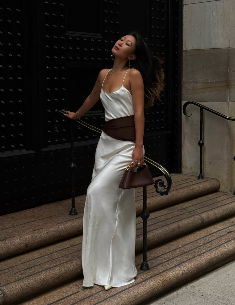 Um vestido acetinado em tom claro destaca-se em um ambiente urbano. A peça, com alças finas e caimento fluido, transmite elegância e sofisticação. A modelo, com cabelo solto, complementa o look com um cinto marrom e uma bolsa estruturada.
