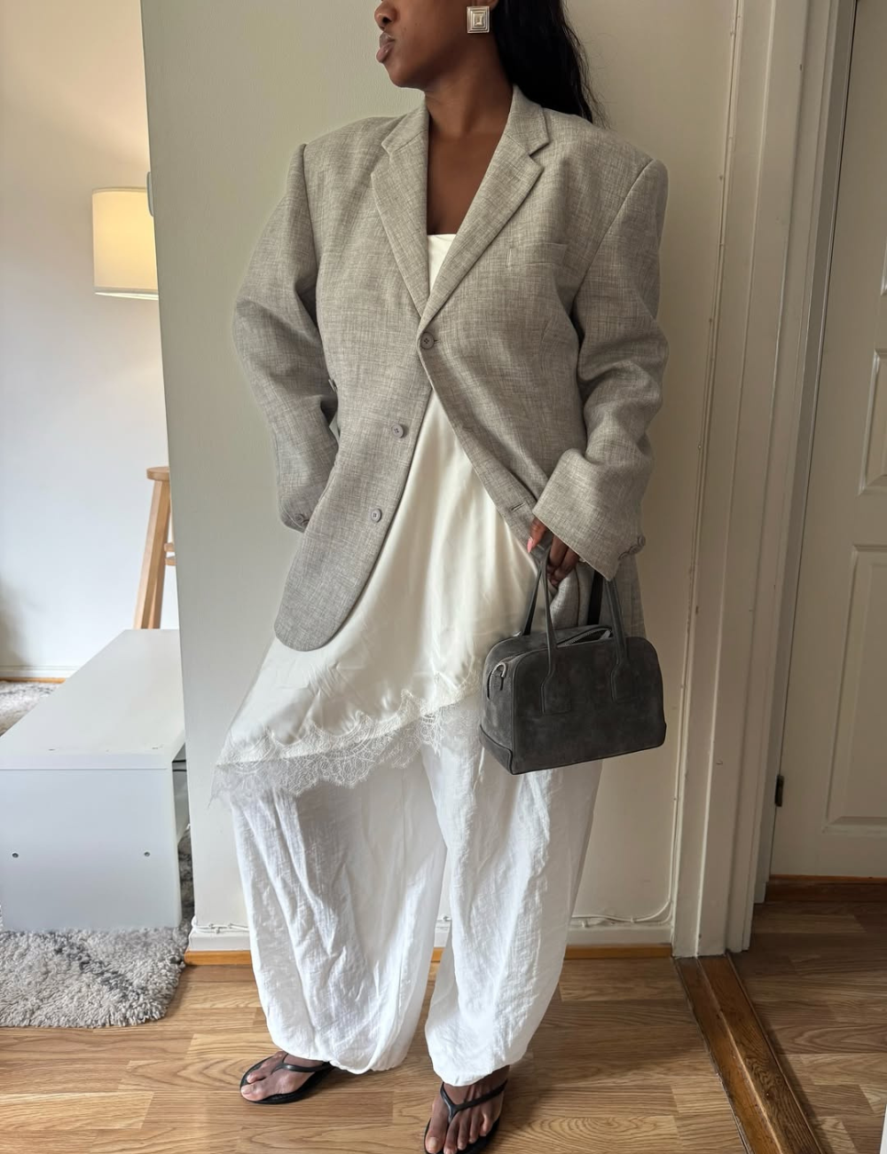 O vestido acetinado, com seu brilho sutil, é complementado por um oversized blazer cinza que traz um toque sofisticado e moderno ao look. A combinação com calças brancas largas e sandálias rasteiras cria um contraste interessante.