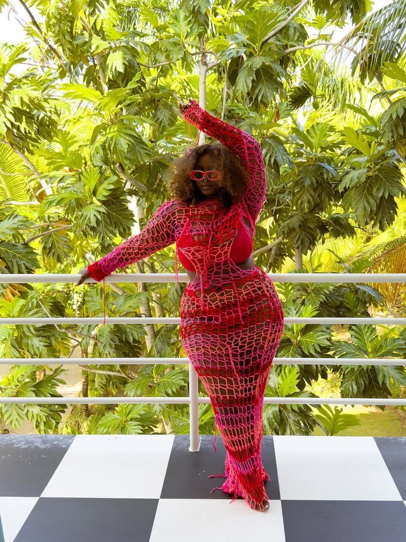 Mulher usando vestido de crochê vermelho vibrante com óculos de sol, em varanda cercada por folhagens tropicais.