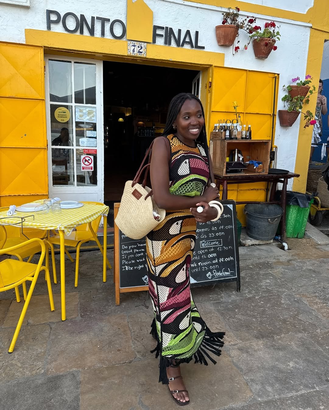 Mulher sorridente com vestido de crochê colorido em frente a restaurante. Ela carrega uma bolsa de palha.
