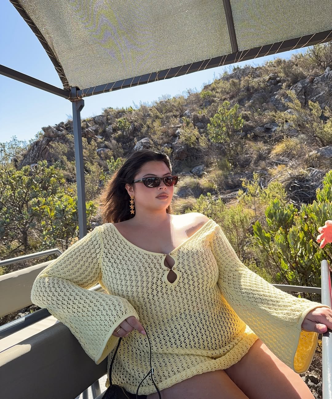 Mulher de óculos escuros, usando vestido de crochê amarelo, sentada em local ensolarado com paisagem montanhosa ao fundo.