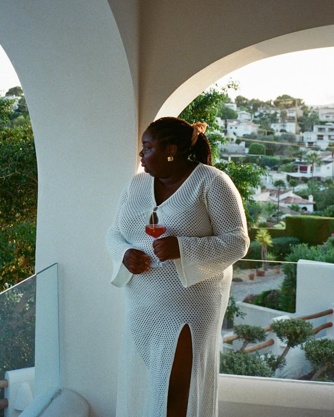 Vestido saída de praia em destaque, confeccionado em material leve e respirável, com design de crochê branco. A peça possui mangas longas e um recorte lateral que valoriza a silhueta. A modelo segura um drinque, complementando o look com elegância.