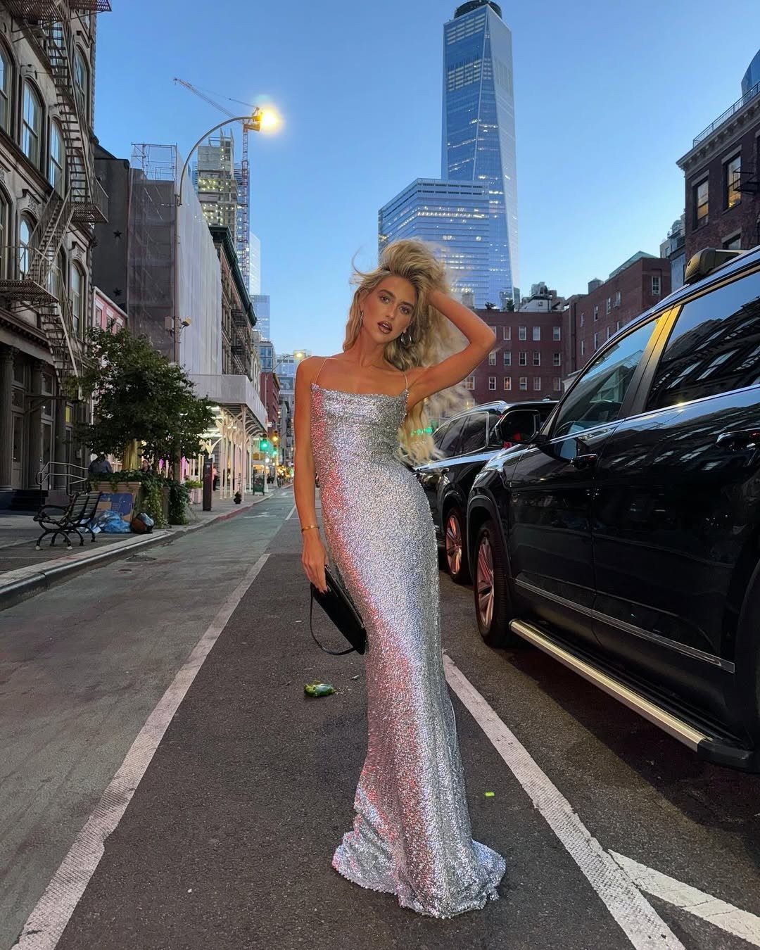 A imagem apresenta uma mulher em uma rua urbana, vestindo um elegante vestido de formatura prateado, com brilho metálico que reflete a luz do ambiente. O modelo é justo, acentuando a silhueta, enquanto ela segura uma bolsa preta.