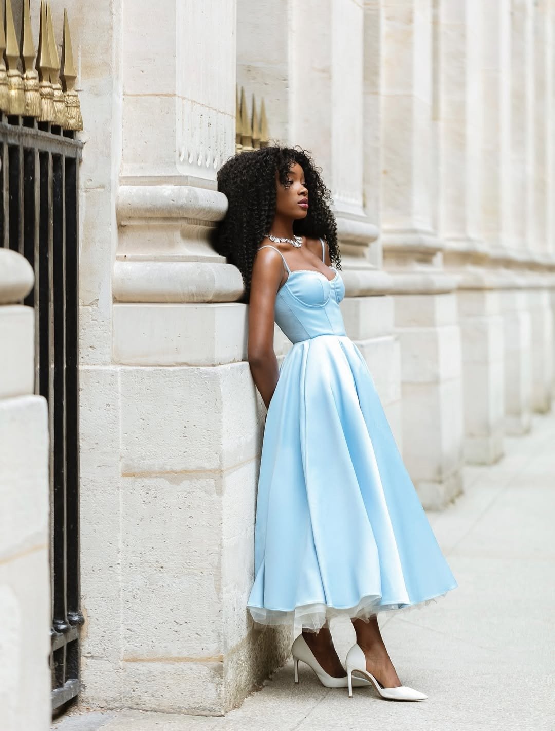 Vestidos de formatura em tom azul claro destaca-se pela elegância e sofisticação. Com um corte midi e detalhes em tule na barra, ele valoriza a silhueta. A modelo usa sapatos brancos de salto alto e um colar delicado, complementando o look.