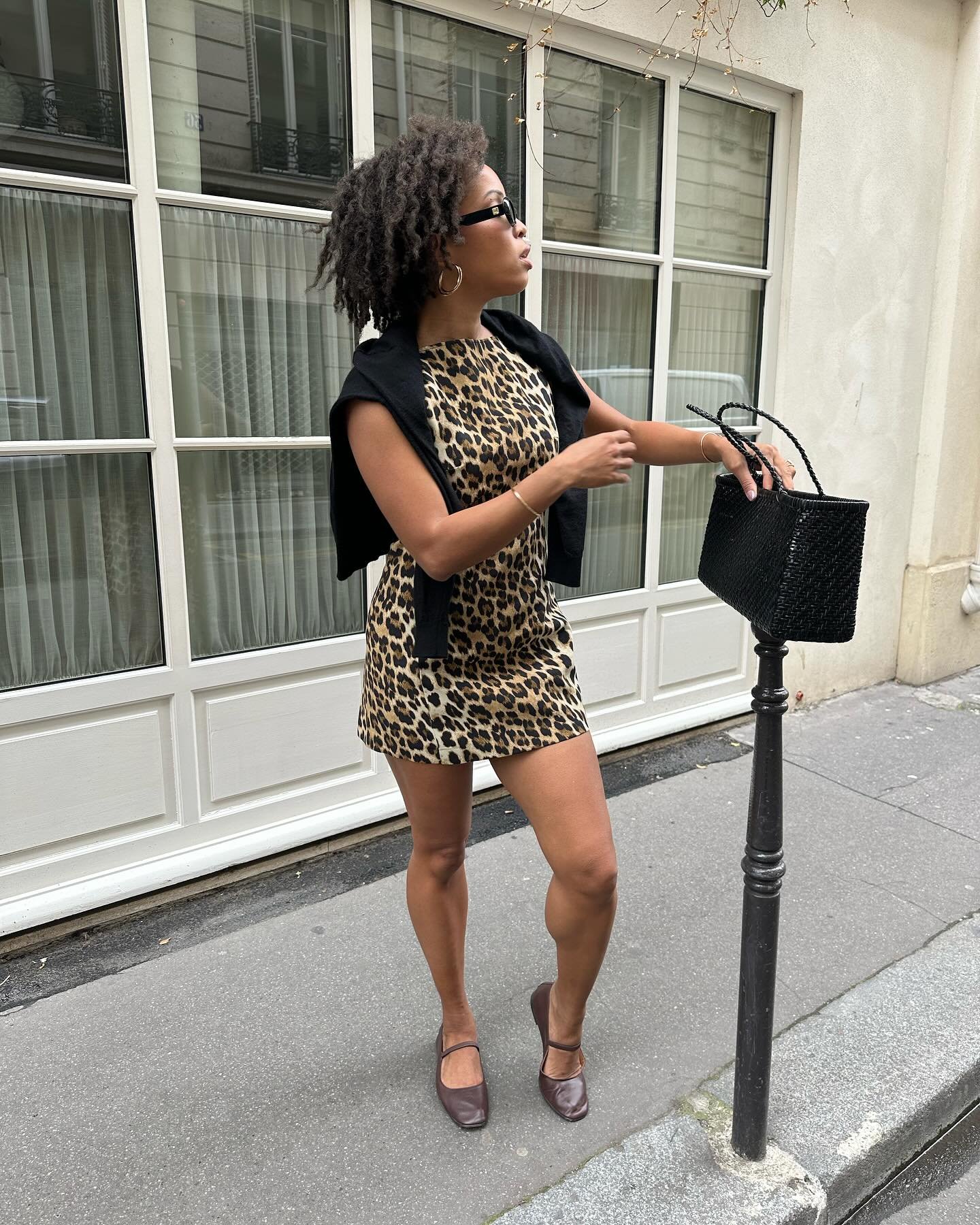 Mulher usando vestido de estampa animal segura bolsa preta na rua junto a uma janela grande.
