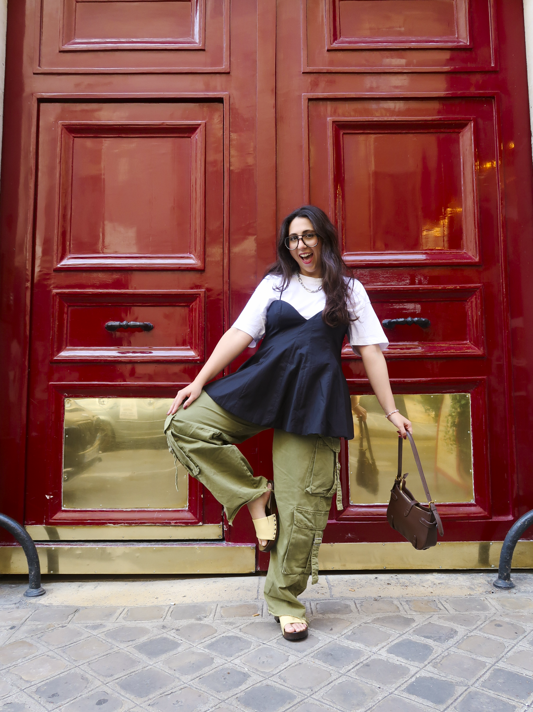 Pessoa sorrindo em frente a uma porta vermelha, usando sapato confortável para viajar e roupa casual estilosa.
