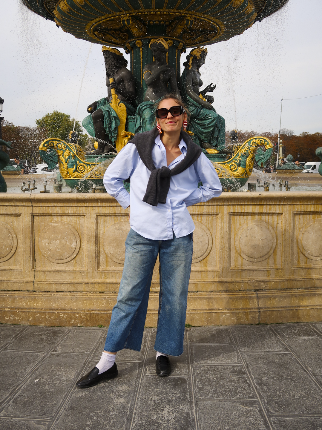 Pessoa com camisa azul e calça jeans diante de uma fonte ornamentada, usando sapato confortável para viajar.