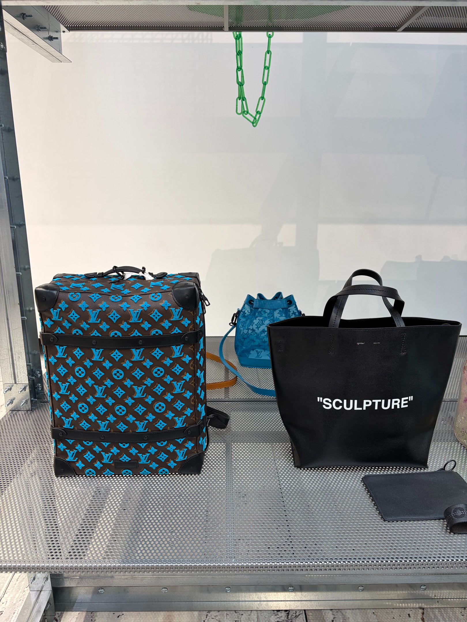 A imagem exibe uma coleção de bolsas de Virgil Abloh, destacando a fusão de streetwear e alta costura. À esquerda, uma mochila em tom marrom com detalhes em azul, apresentando o icônico monograma da Louis Vuitton. À direita, uma tote preta com a palavra 