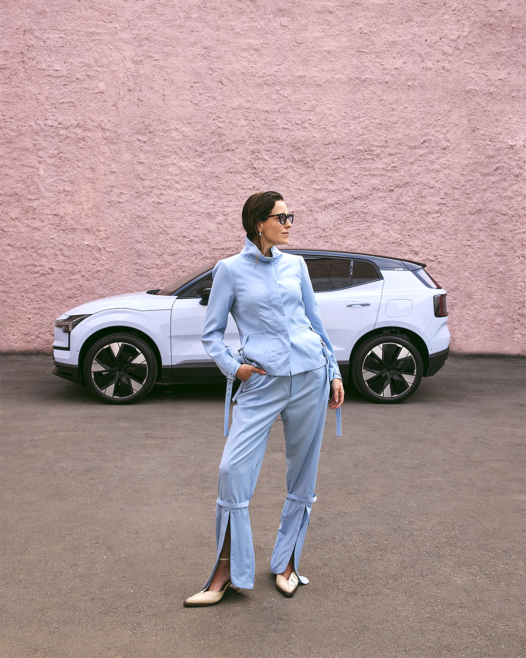 Usuária de óculos escuros veste azul claro elegante, em frente a um Volvo branco, com fundo rosa.