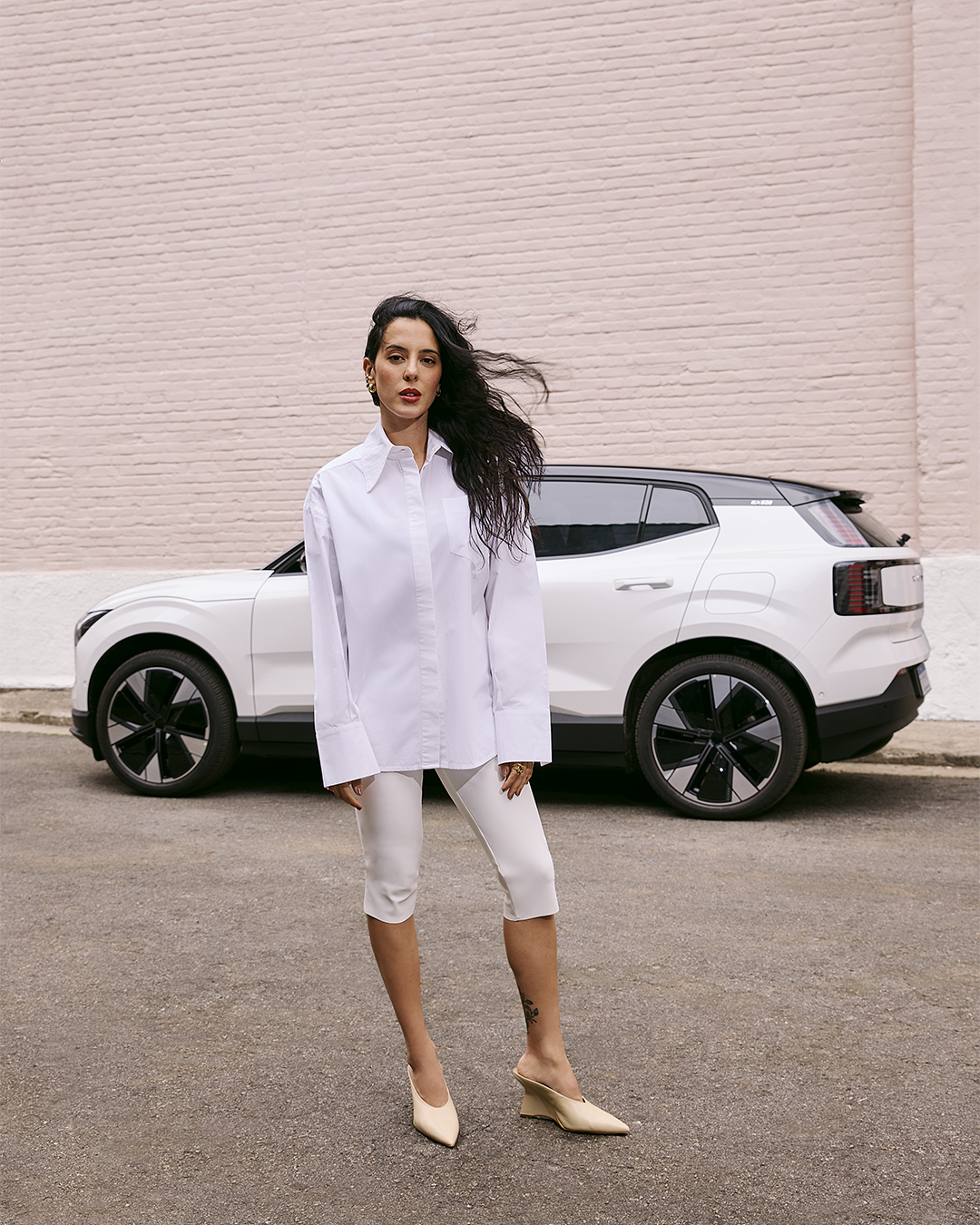 Pessoa com roupa branca em frente a um carro branco da Volvo estacionado, parede rosa ao fundo.