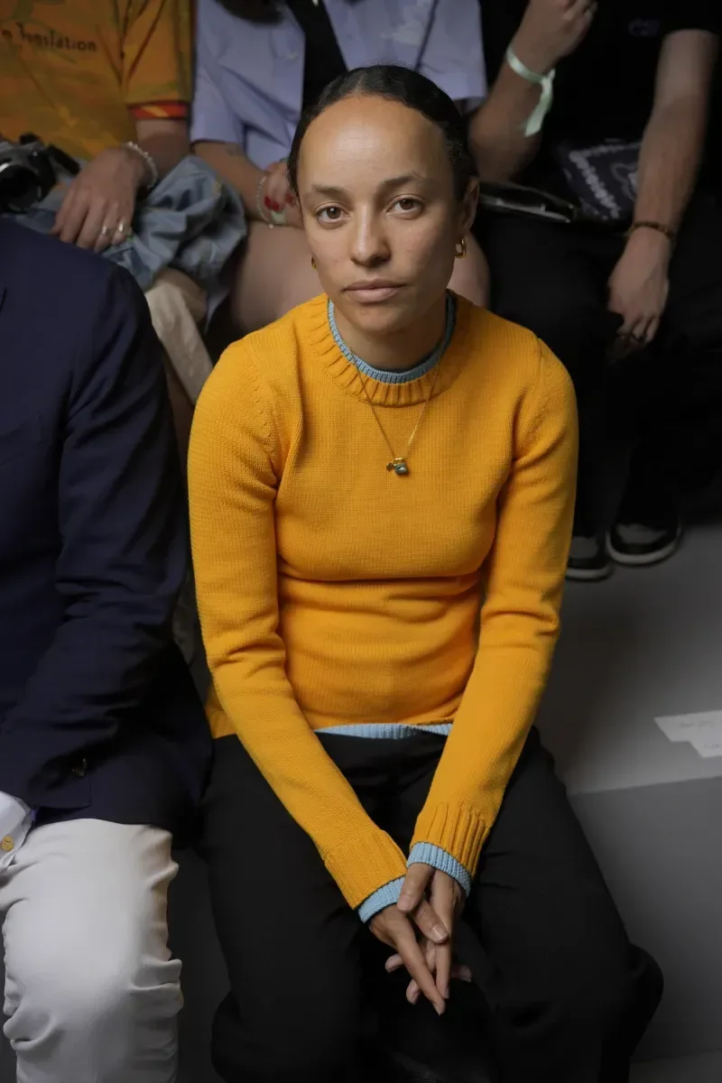 Grace Wales Bonner aparece sentada em um ambiente de moda, usando um suéter amarelo vibrante com detalhes em azul claro, que destaca seu estilo contemporâneo e minimalista.