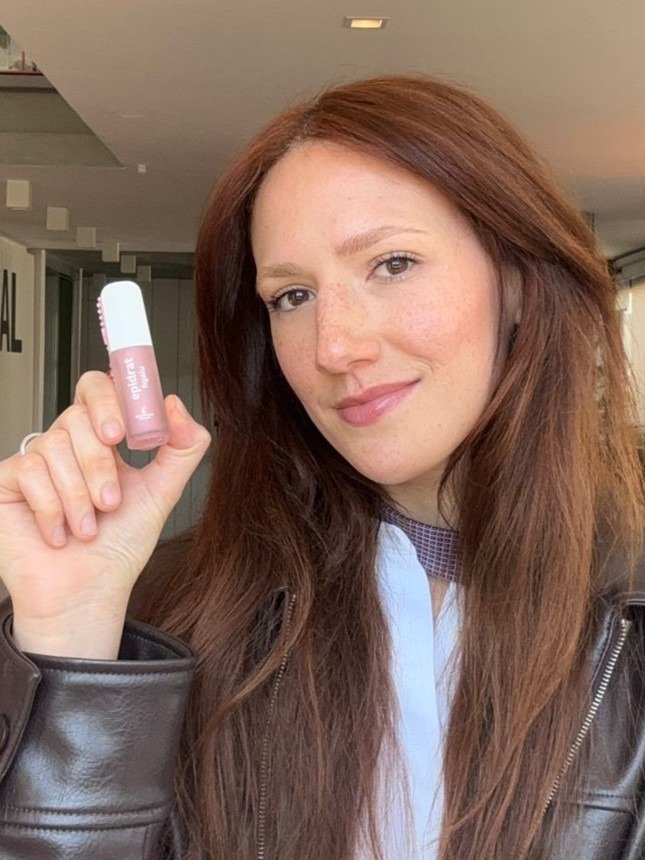 A imagem apresenta uma mulher com cabelo longo e castanho, segurando um Lip Oil Epidrat Hyalu em tom rosa suave, um lip oil hidratante e com proteção solar