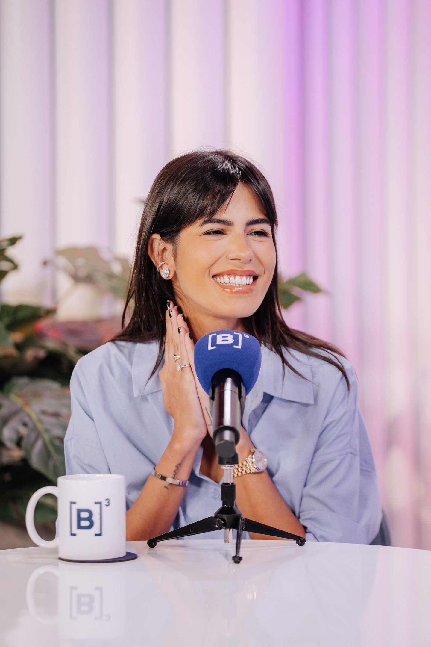 Anttónia exibe um sorriso radiante em um ambiente moderno, com iluminação suave em tons de rosa. Ela veste uma camisa azul clara, que destaca sua elegância e estilo. Os acessórios, como os brincos grandes e anéis delicados, complementam seu visual.