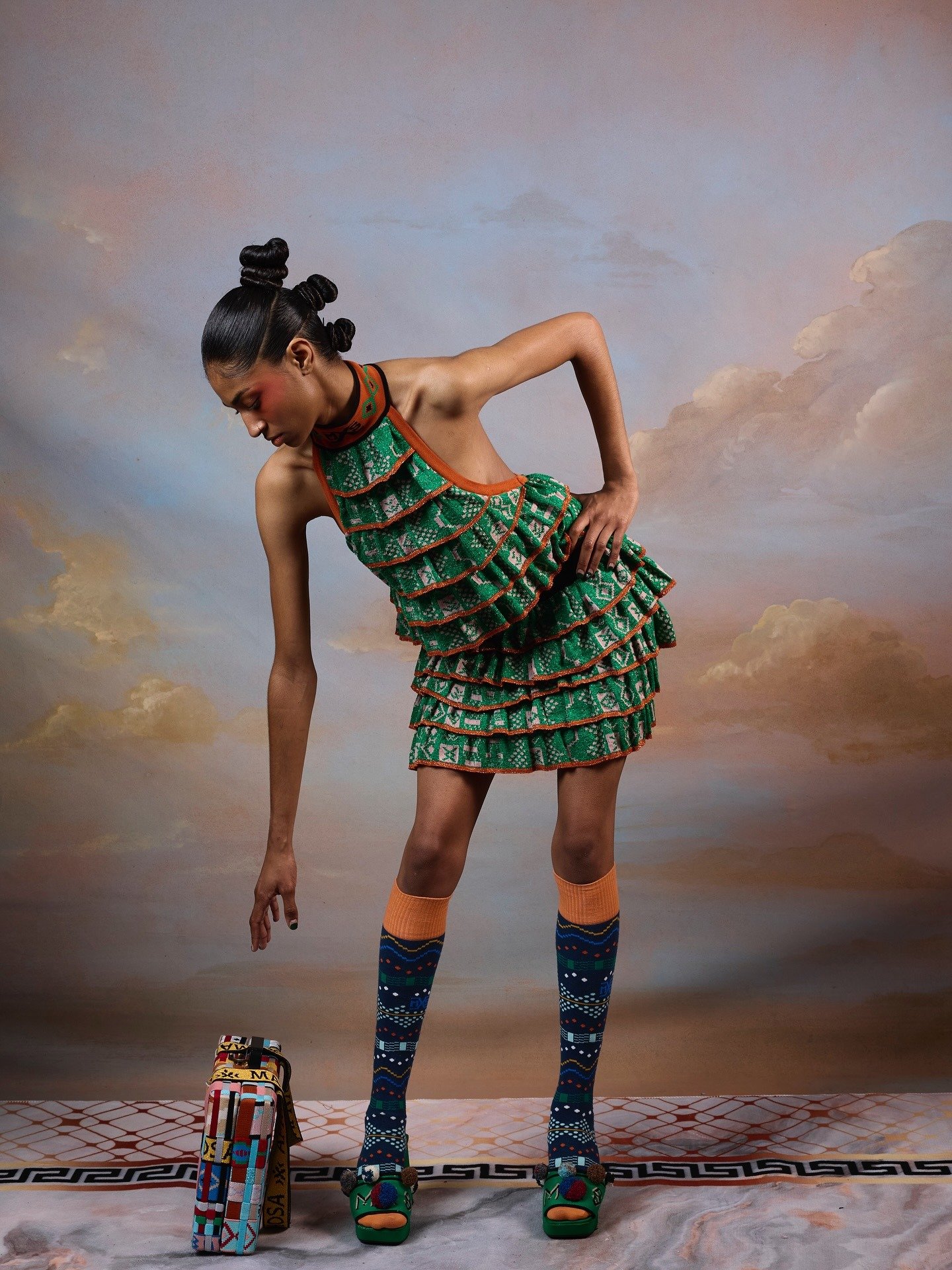 Modelo posa inclinada com vestido verde de babados geométricos, meias coloridas e sandálias verdes, evocando afrofuturismo.
