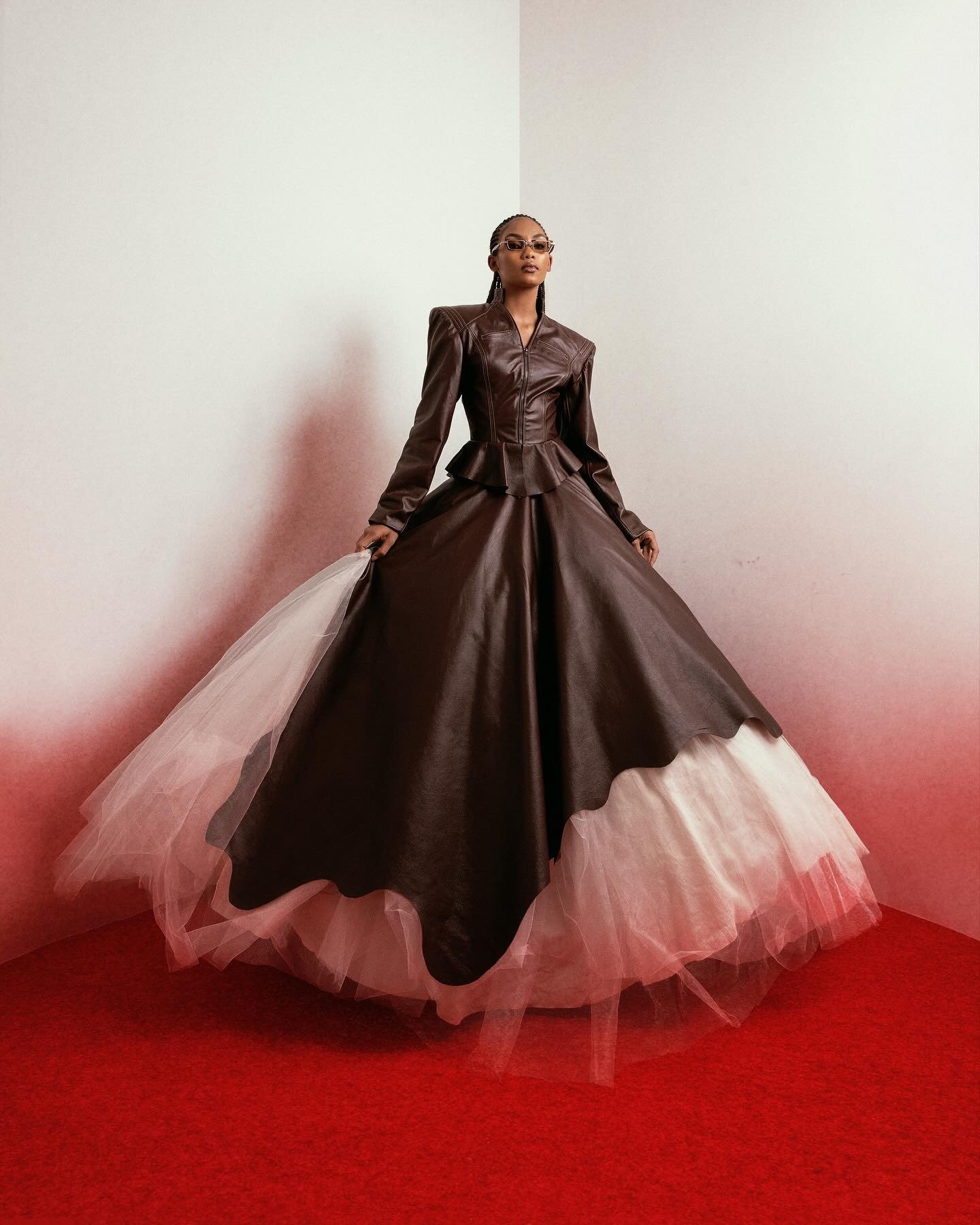 Modelo posa com vestido de afrofuturismo de couro marrom estruturado e saia ampla com tule branco, em cenário minimalista.