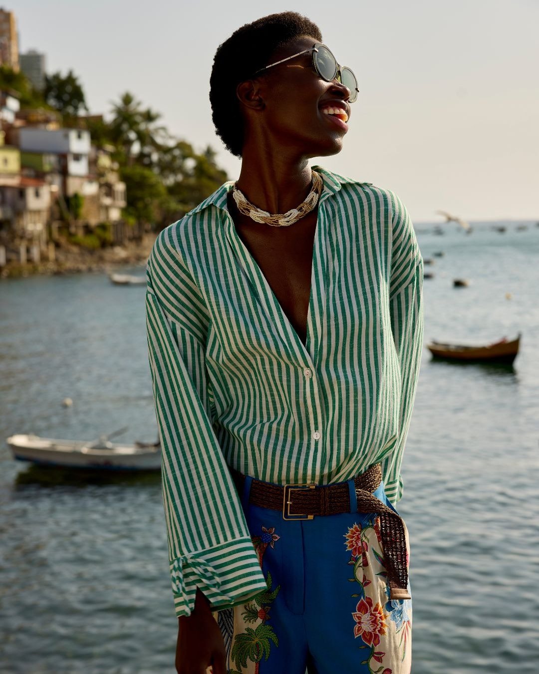 Modelo posa sorrindo à beira-mar com camisa listrada verde e branca, shorts azul vibrante com bordados, a cara do alto verão