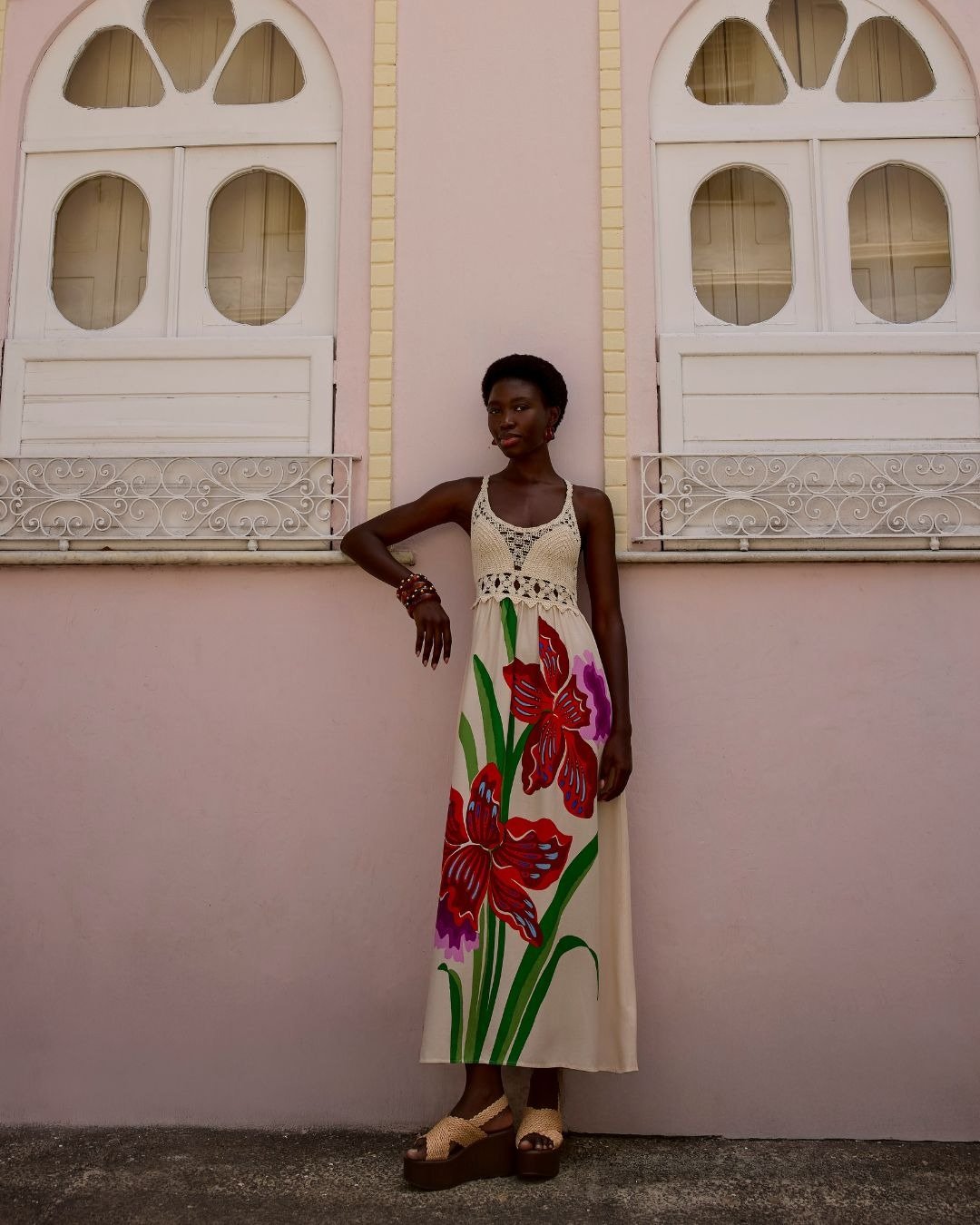 Modelo posa com vestido longo de tecido leve com estampa floral vermelha e lilás, top de crochê cru e sandálias plataforma,