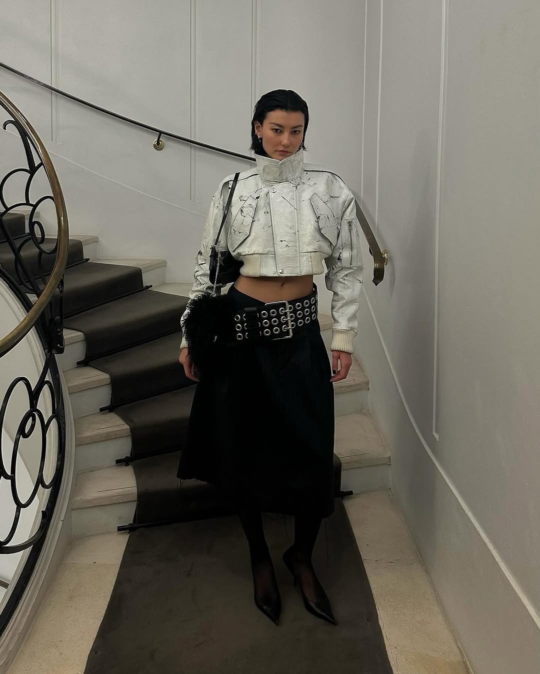 Modelo posa com jaqueta cropped branca de couro texturizado e gola funil alta, combinada a saia preta midi, cinto largo com