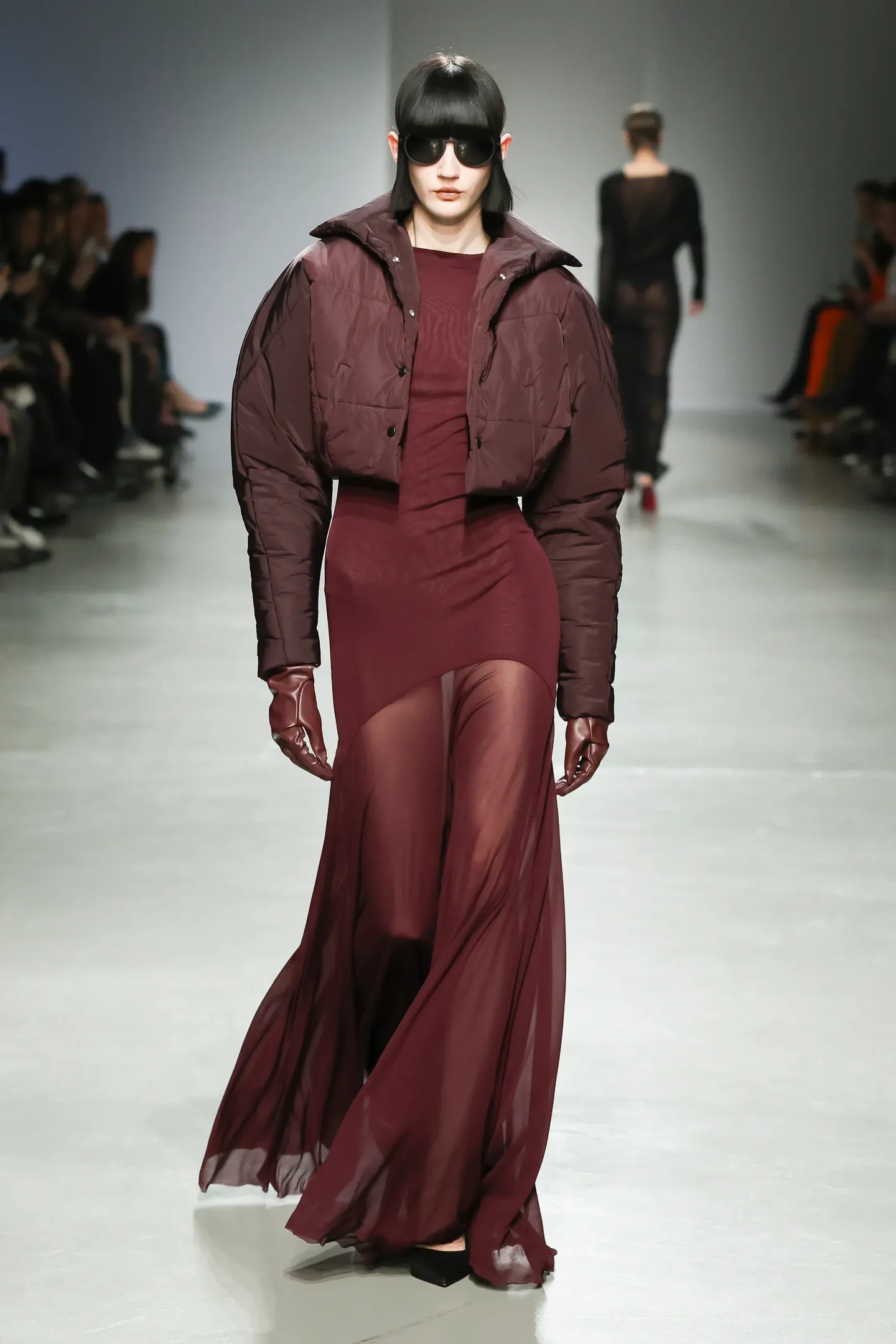 Modelo desfila com vestido longo em tom bordô, com parte superior transparente e jaqueta acolchoada.