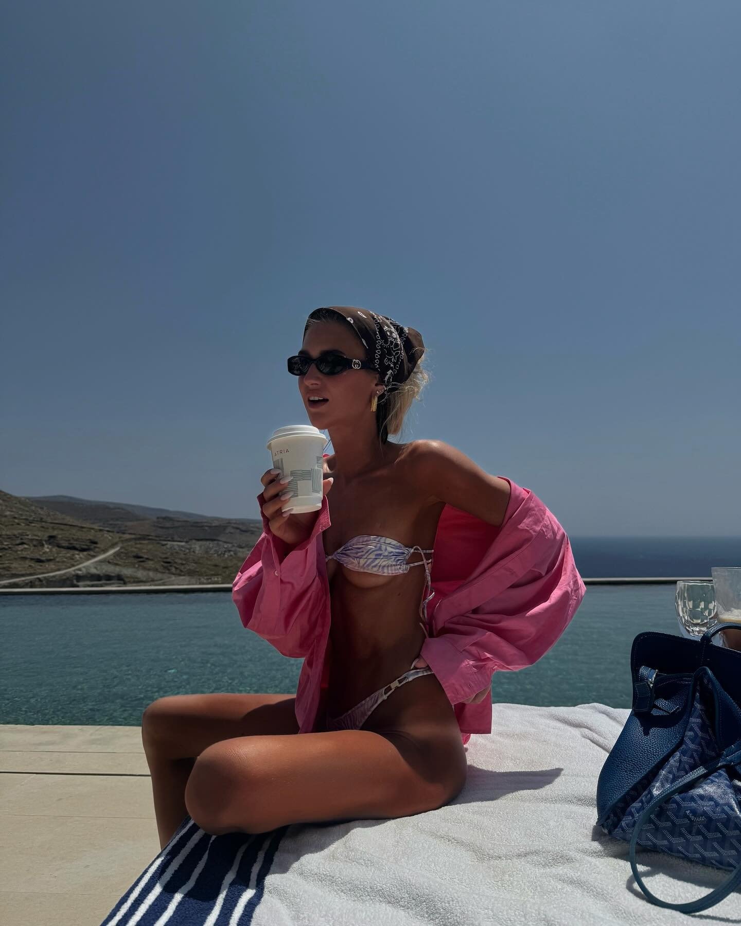 Modelo senta à beira da piscina com looks com bandana marrom estampada, biquíni lilás e camisa rosa oversized