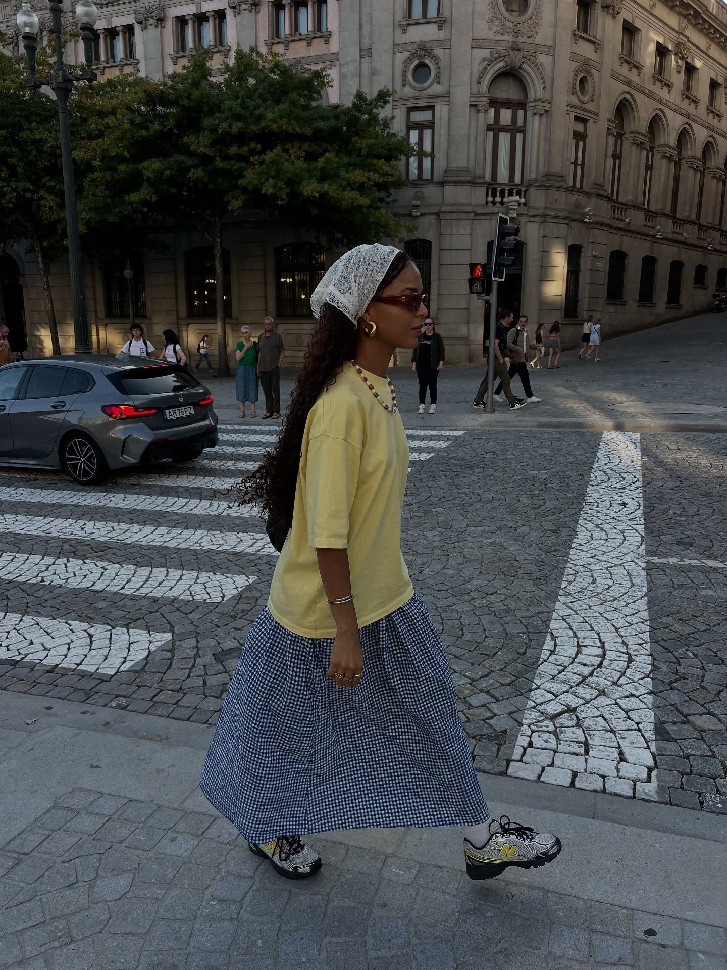 Modelo caminha com looks com bandana branca de renda, camiseta amarela oversized, saia midi xadrez azul e tênis esportivo.
